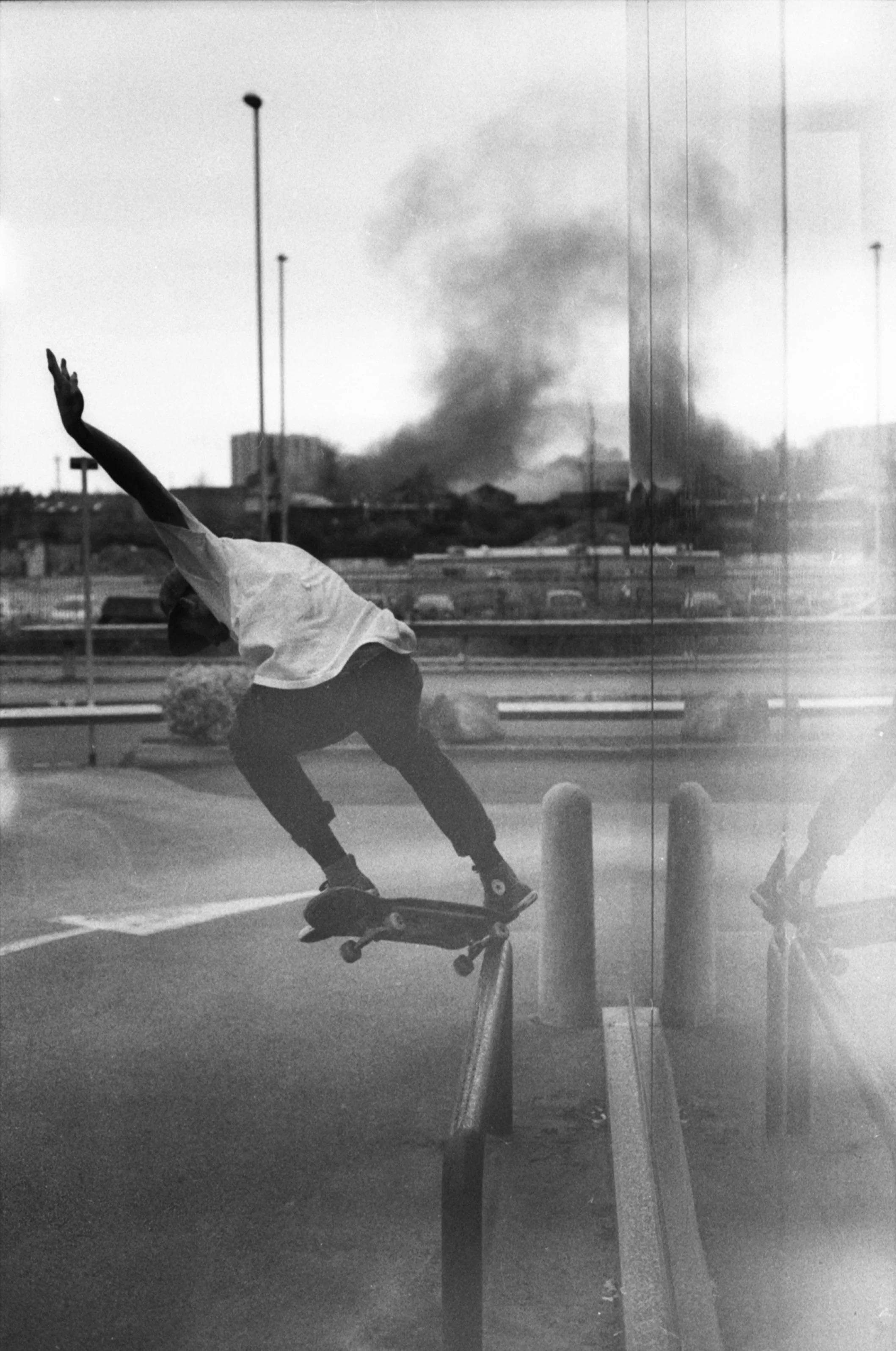 A person skateboarding on a rail at a skatepark with smoke rising in the background, viewed through a glass surface.