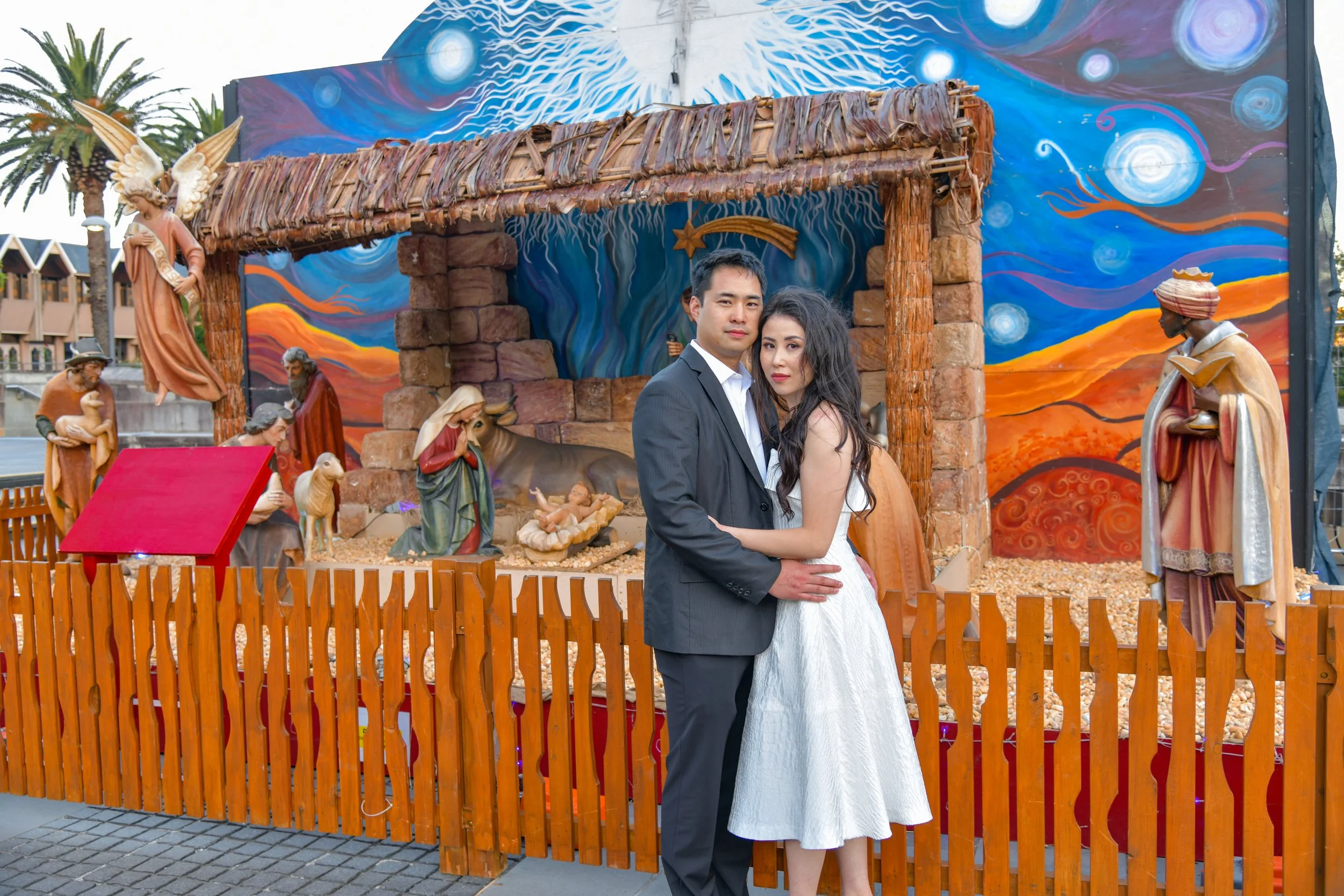 A couple dressed in formal attire, standing close in front of a nativity scene featuring figures of Mary, Joseph, baby Jesus, and animals, set against a colorful painted backdrop.