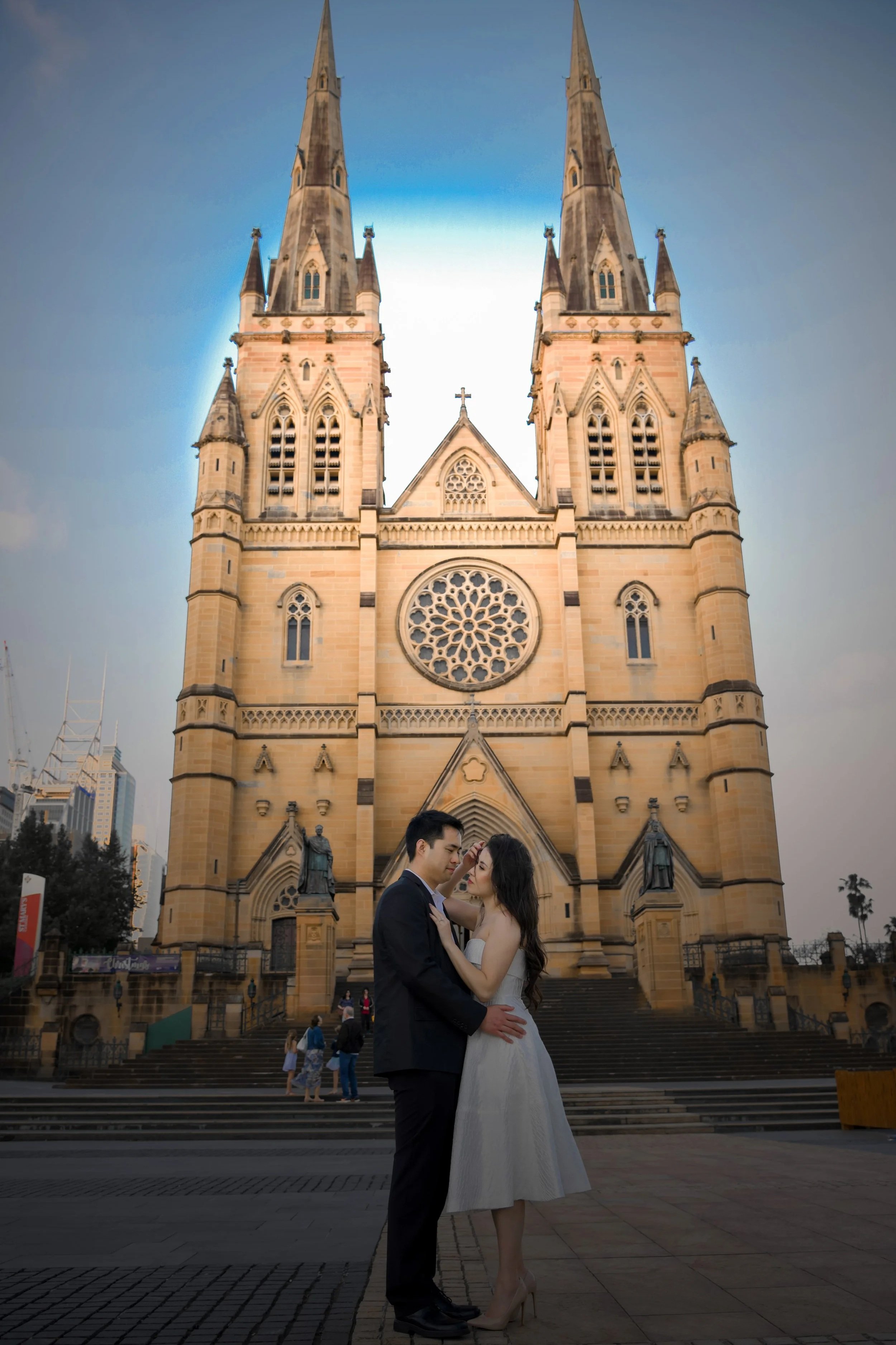 A couple in wedding attire stands close together in front of St. Mary's Basilica, a Gothic-style church with tall spires and detailed stonework, in a cityscape setting during dusk.