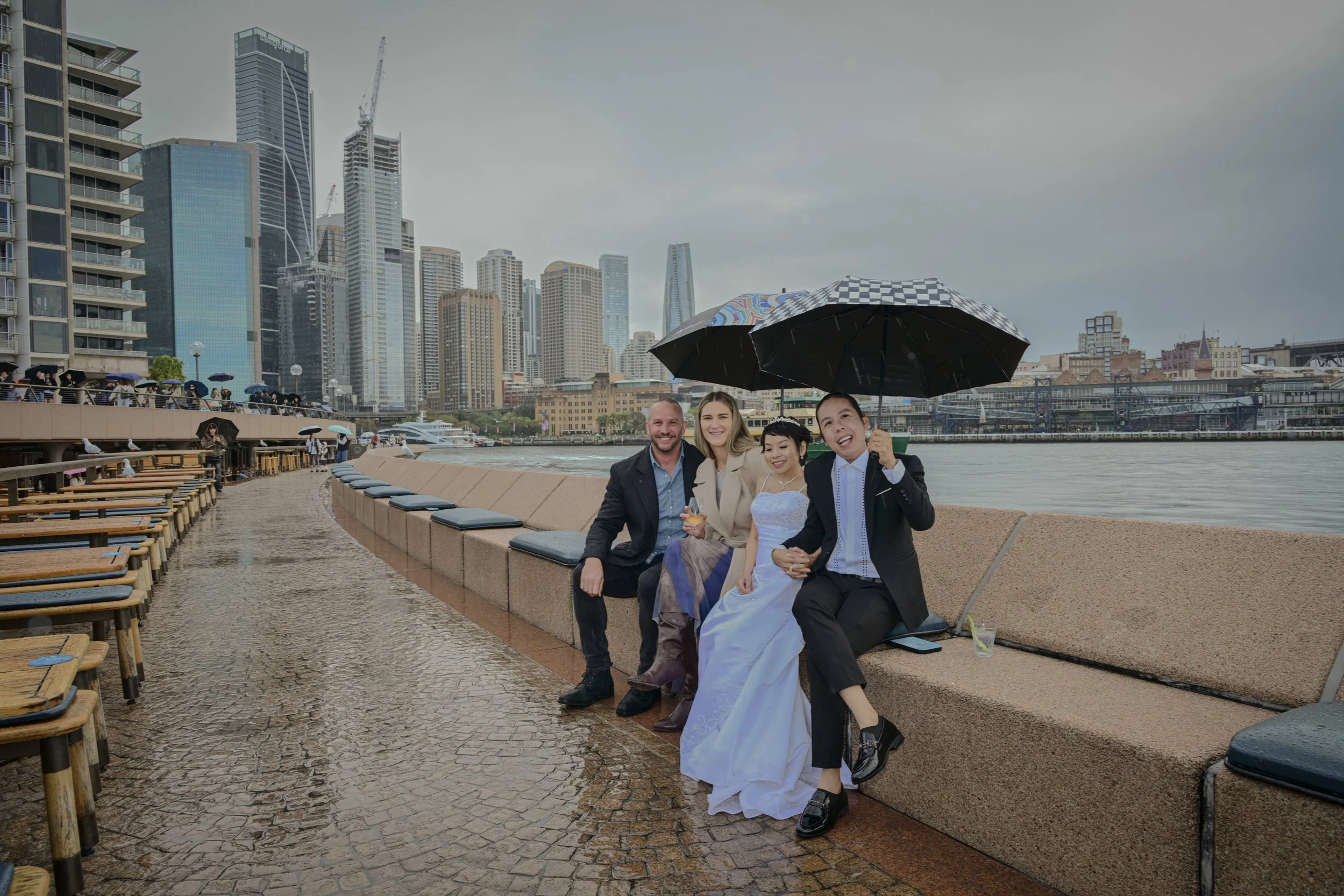People sitting on a bench by the river with city skyscrapers in the background, holding umbrellas on a rainy day.