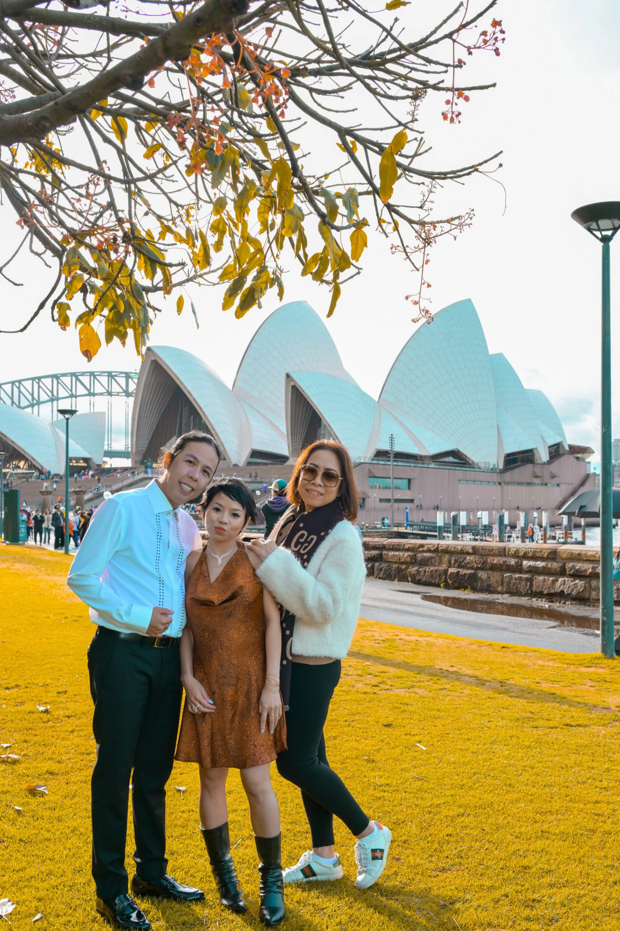 Three women standing together outdoors in front of the Sydney Opera House, with a tree near the top of the image and a streetlamp on the right.