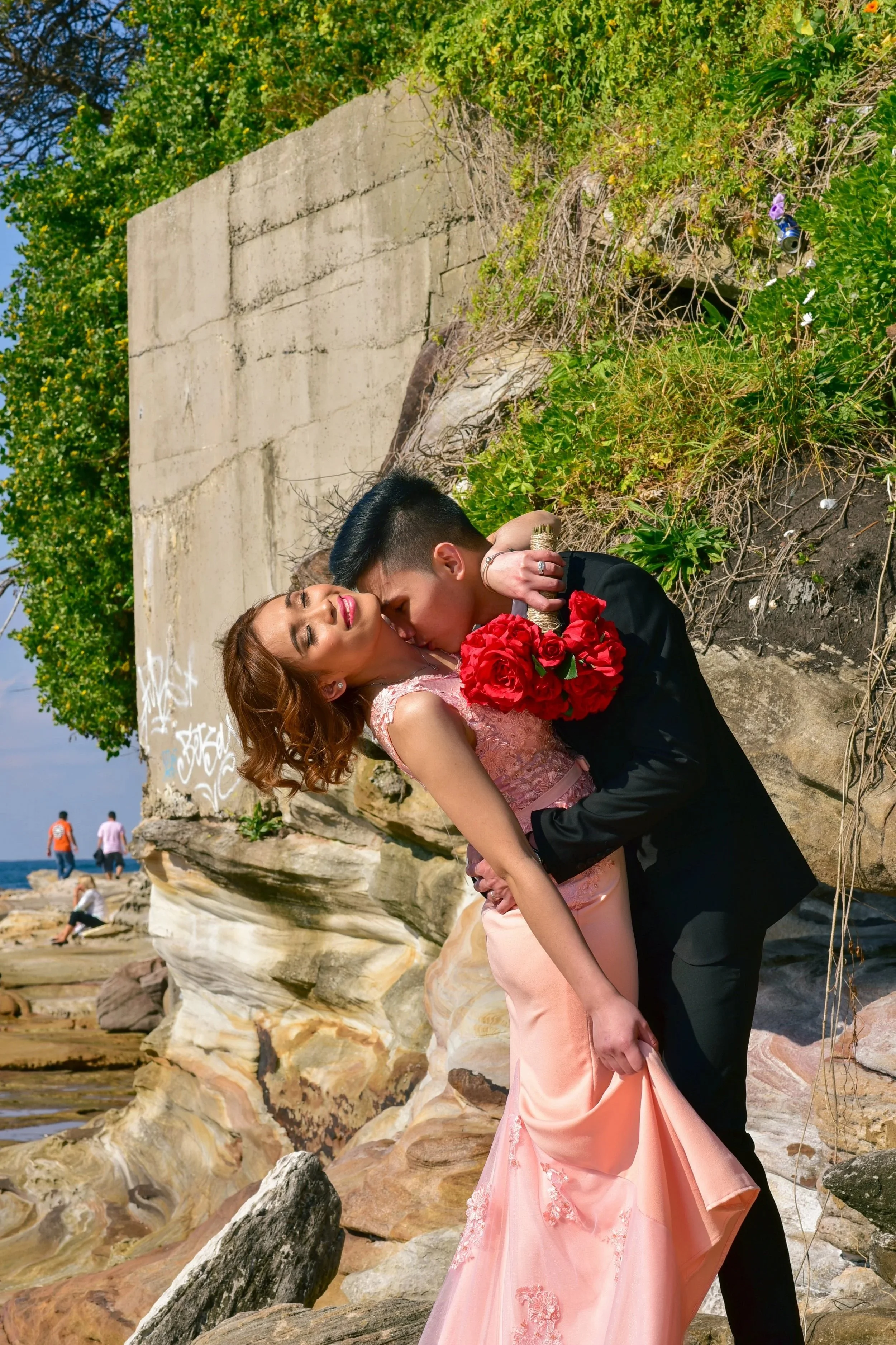 A couple, dressed in formal attire, sharing a romantic kiss on a rocky beach, with the man holding a bouquet of red roses and the woman wearing a pink gown, under a concrete wall covered with greenery.