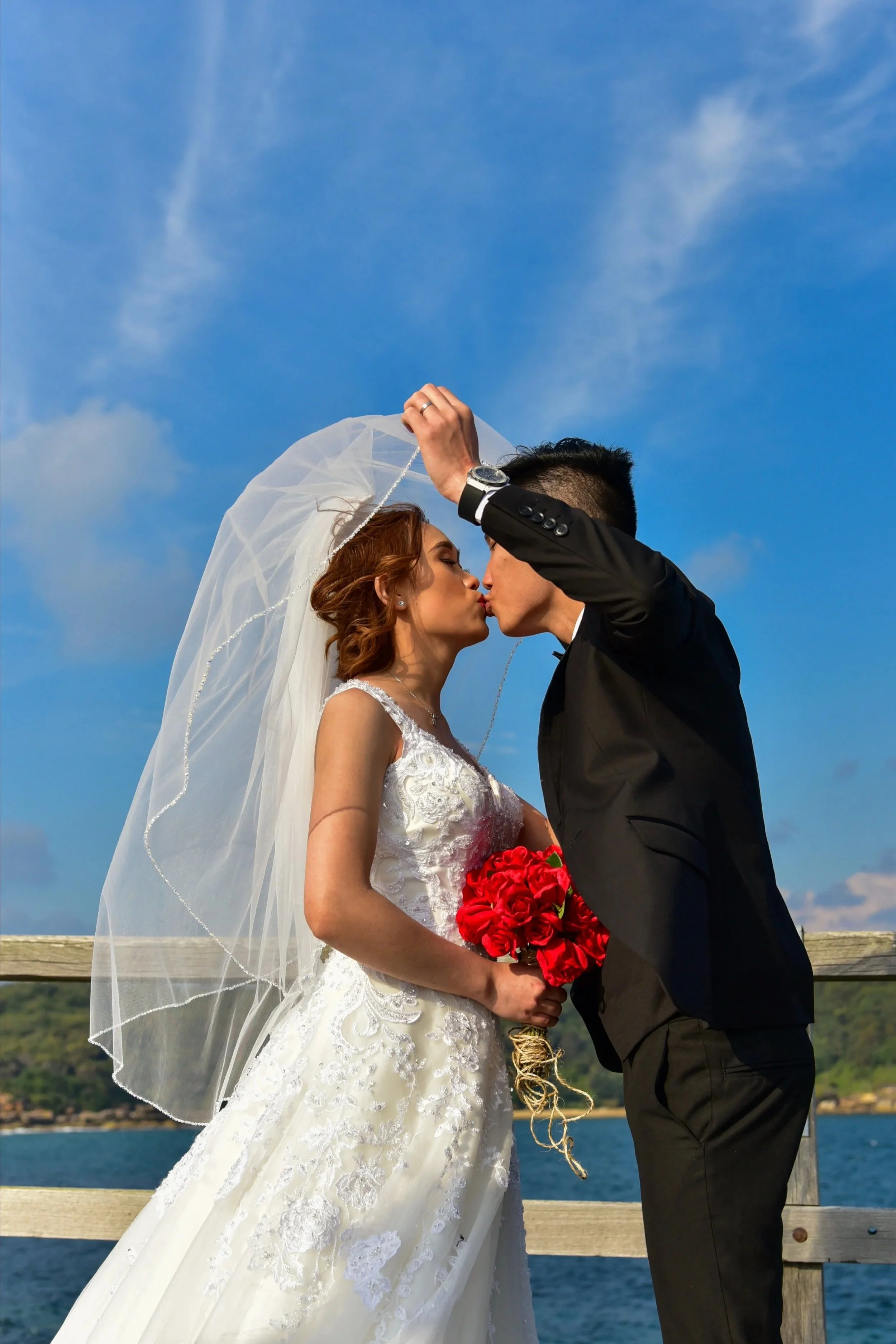 A bride and groom share a kiss outdoors on a wooden dock with water and hills in the background, under a partly cloudy sky.