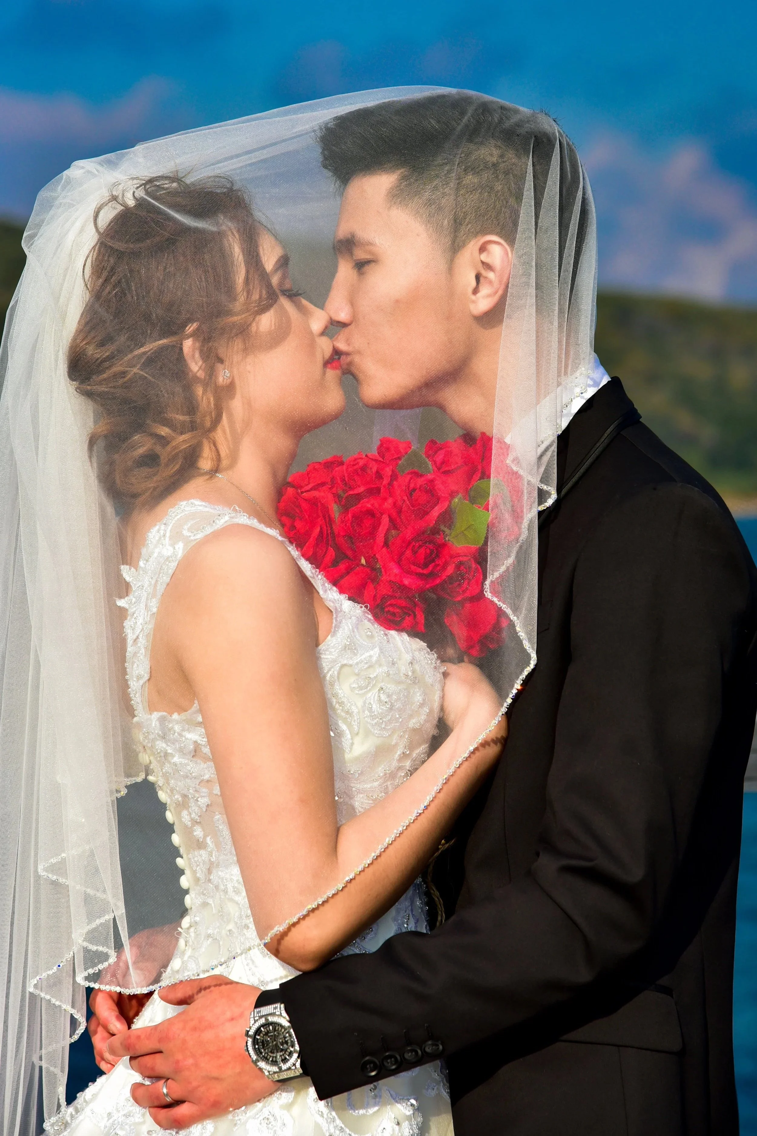 A bride and groom kiss under a wedding veil, with the bride holding a bouquet of red roses, outdoors with a blue sky background.