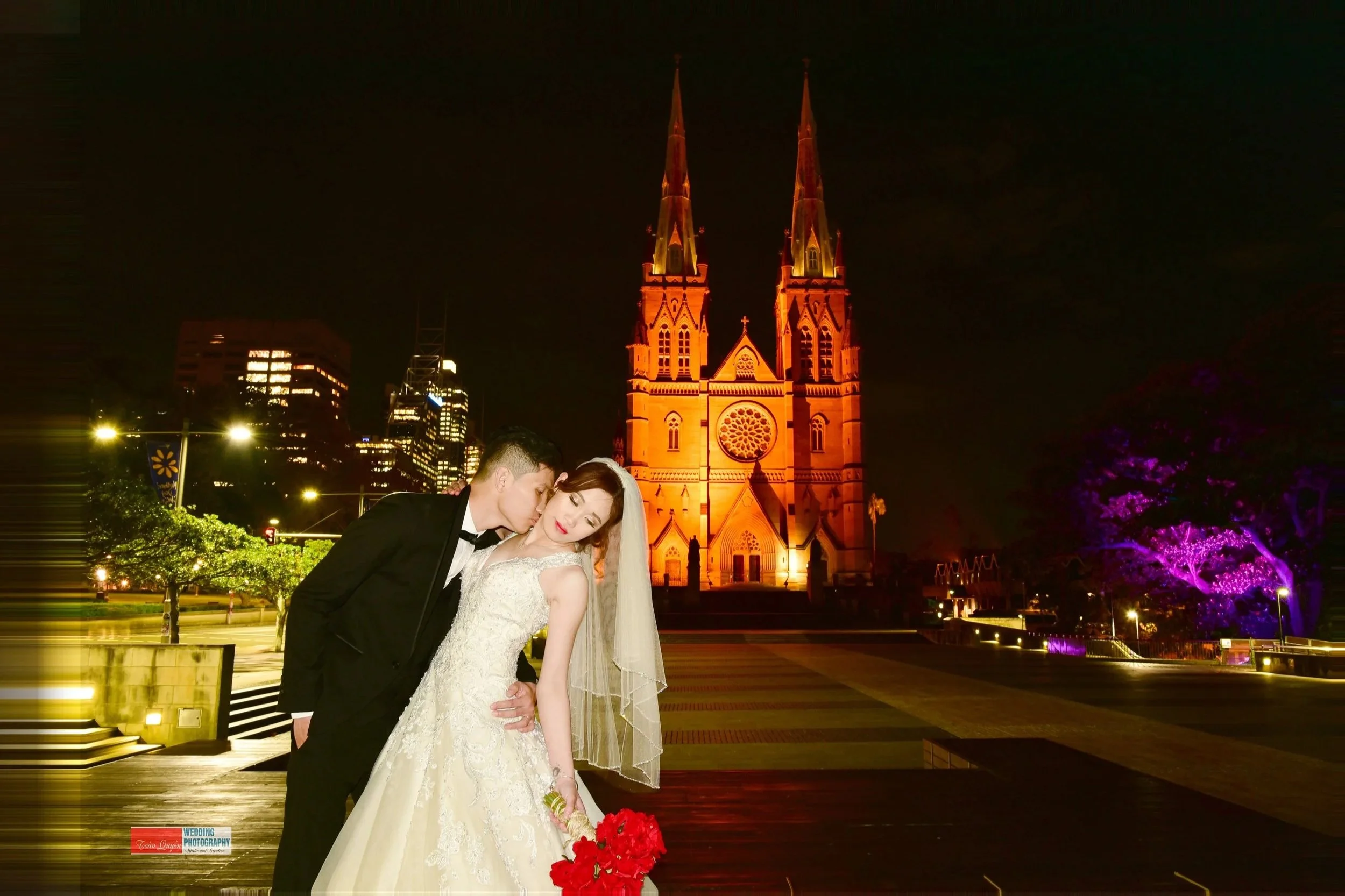 A newlywed couple, with the groom kissing the bride on her cheek, stands in front of a illuminated cathedral at night. The bride is holding a bouquet of red flowers, and the scene is set in an urban area with city lights and buildings in the backgrou