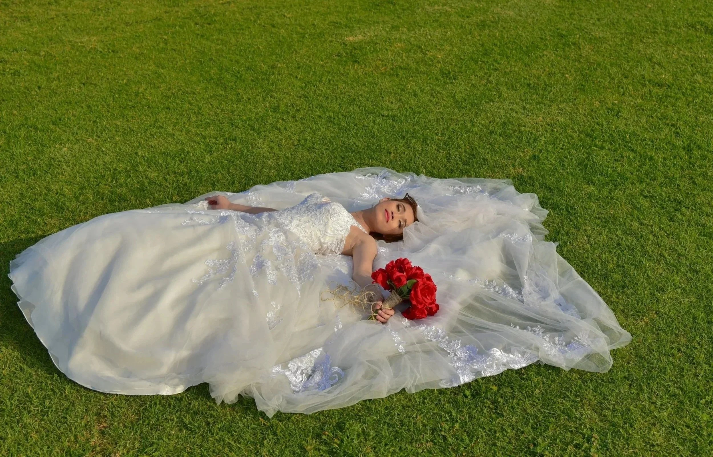 A bride lying on a grassy field in her wedding dress, holding a bouquet of red roses.