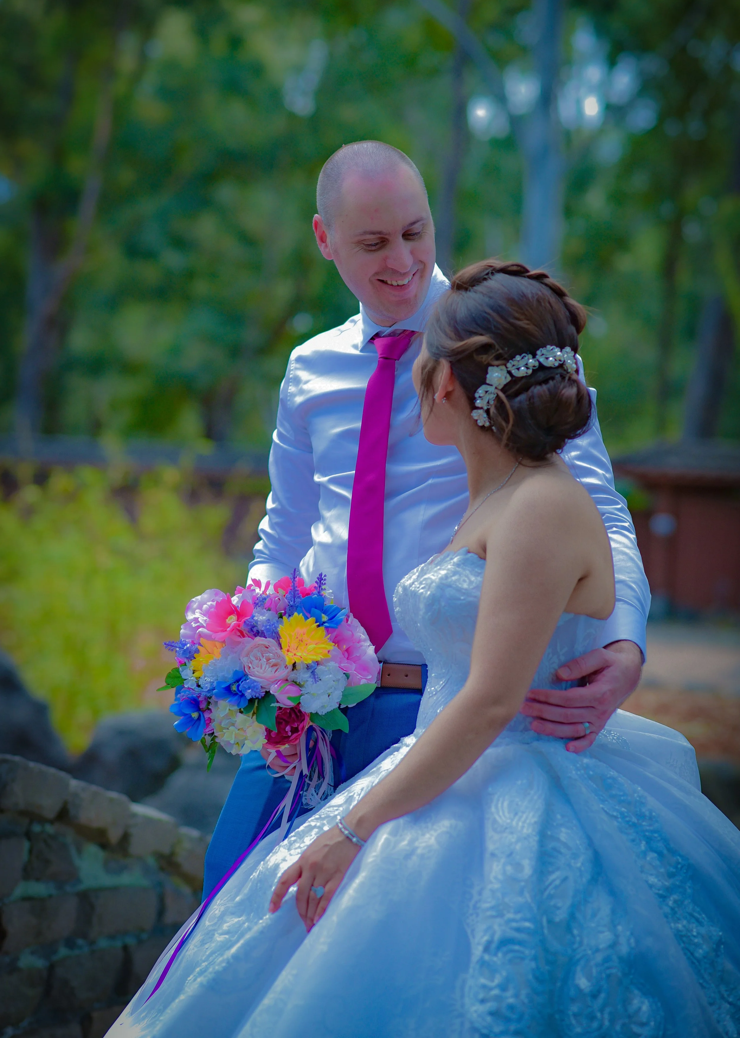 A bride and groom smiling at each other outdoors with a green background, the bride is in a white wedding dress with a floral hairpiece, and the groom is in a white shirt with a purple tie, holding a colorful bouquet.