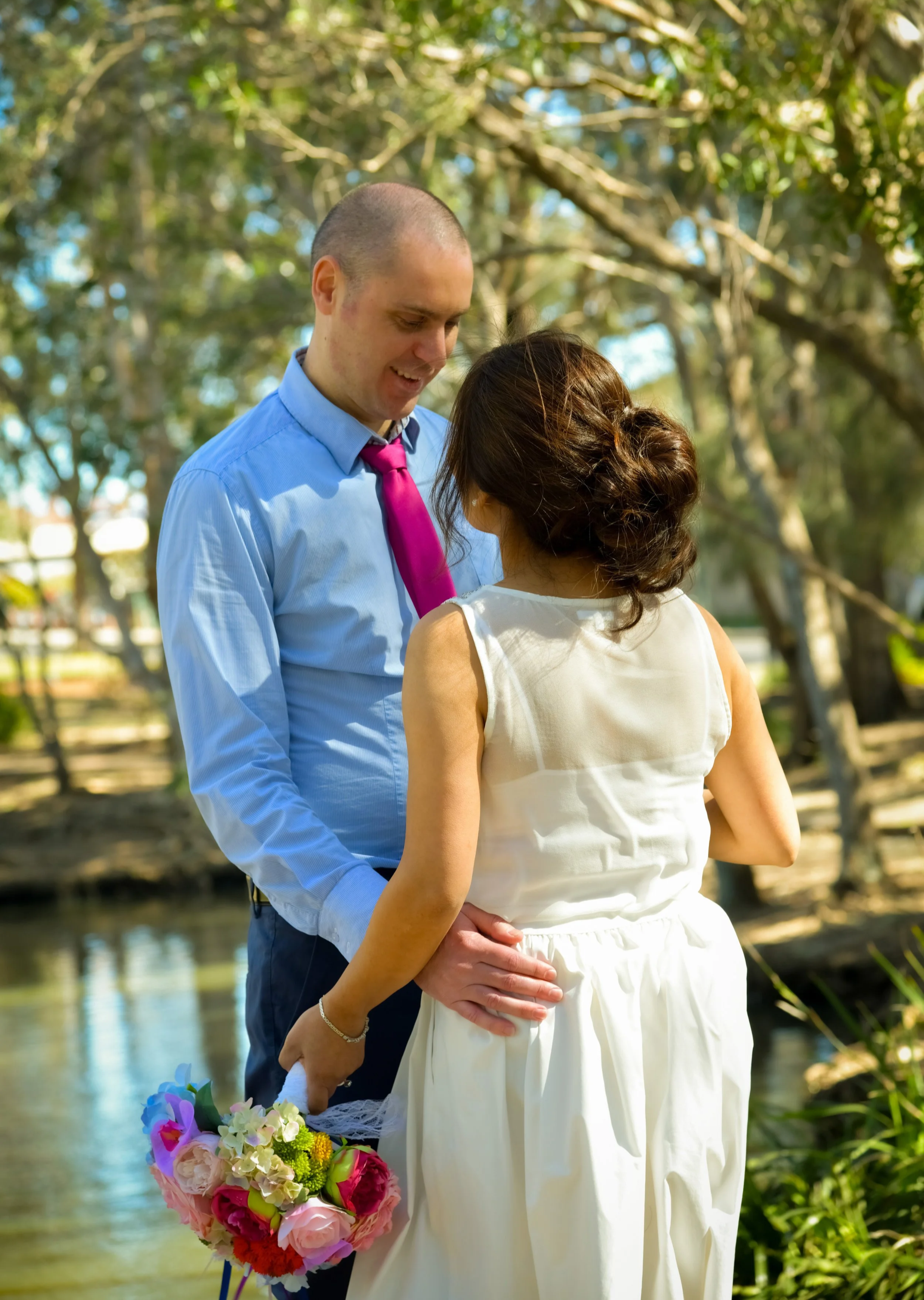 A couple dressed in formal attire, holding hands, standing outdoors by a river, with trees in the background. The woman is holding a colorful bouquet of flowers.
