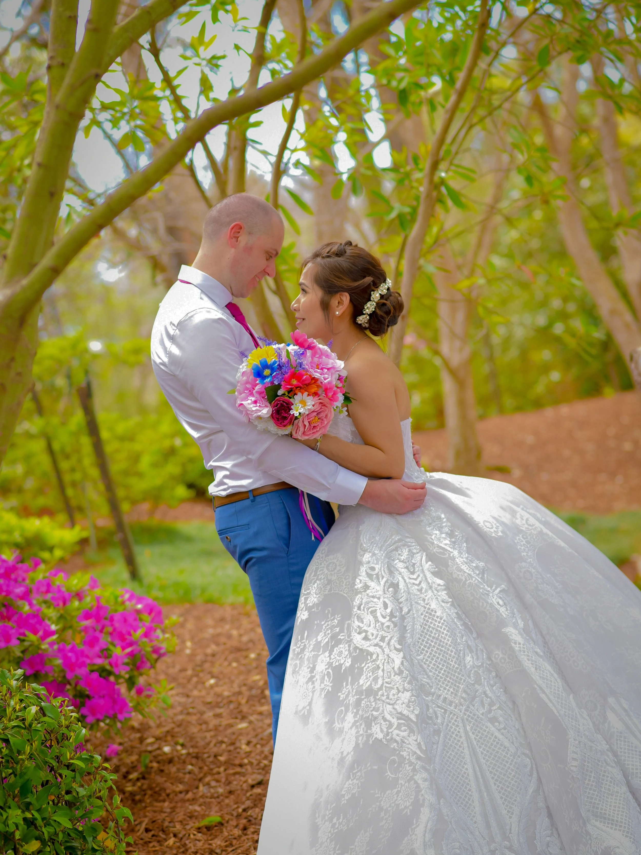 A newlywed couple stands close together outdoors in a garden, with the groom lifting the bride, both smiling and looking lovingly at each other. The bride is holding a bouquet of colorful flowers, and they are surrounded by trees and pink flowers.