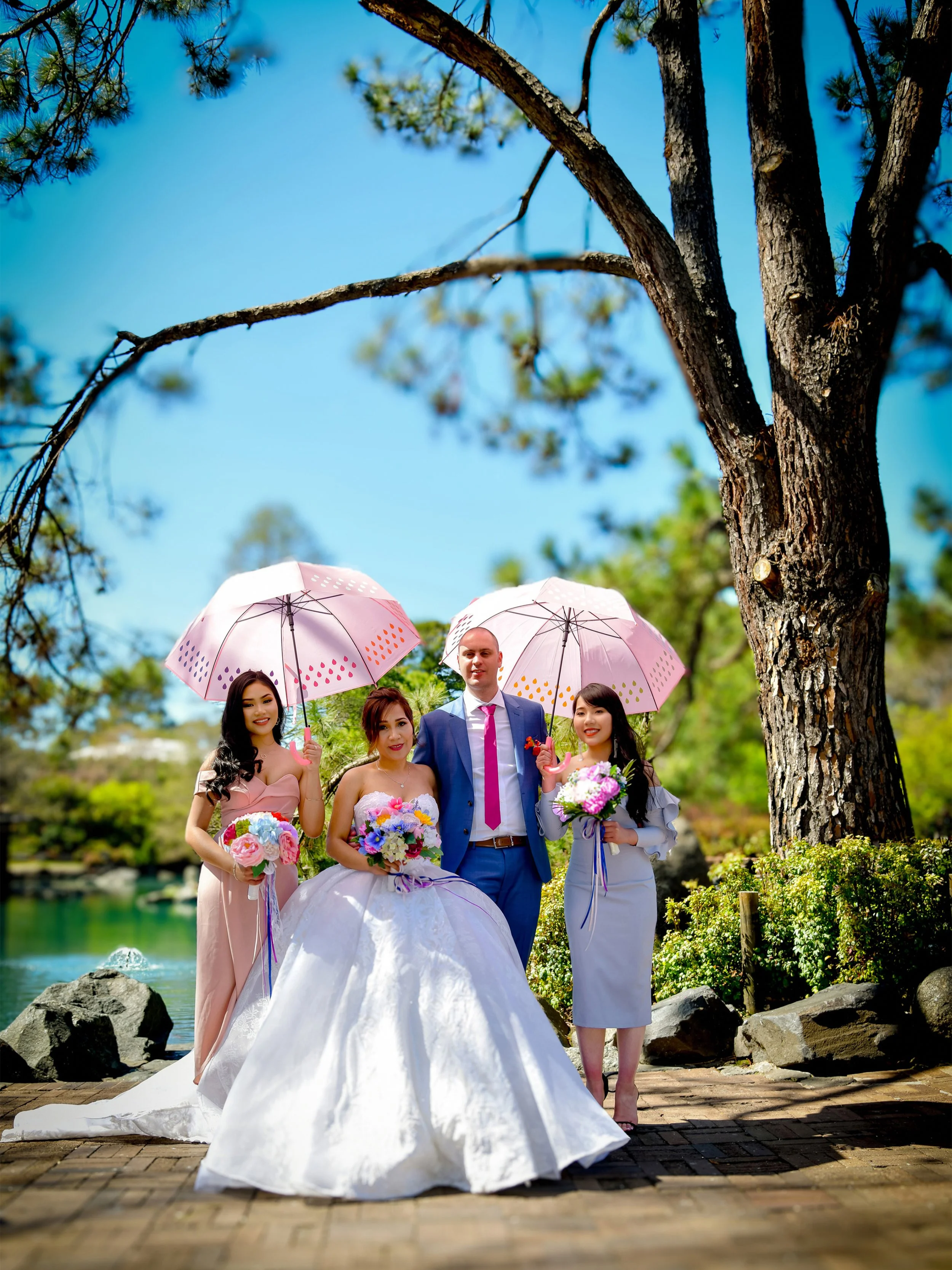 A bride in a white gown and three bridesmaids in pastel dresses holding bouquets, standing outdoors by a pond with a large tree overhead, holding pink umbrellas.