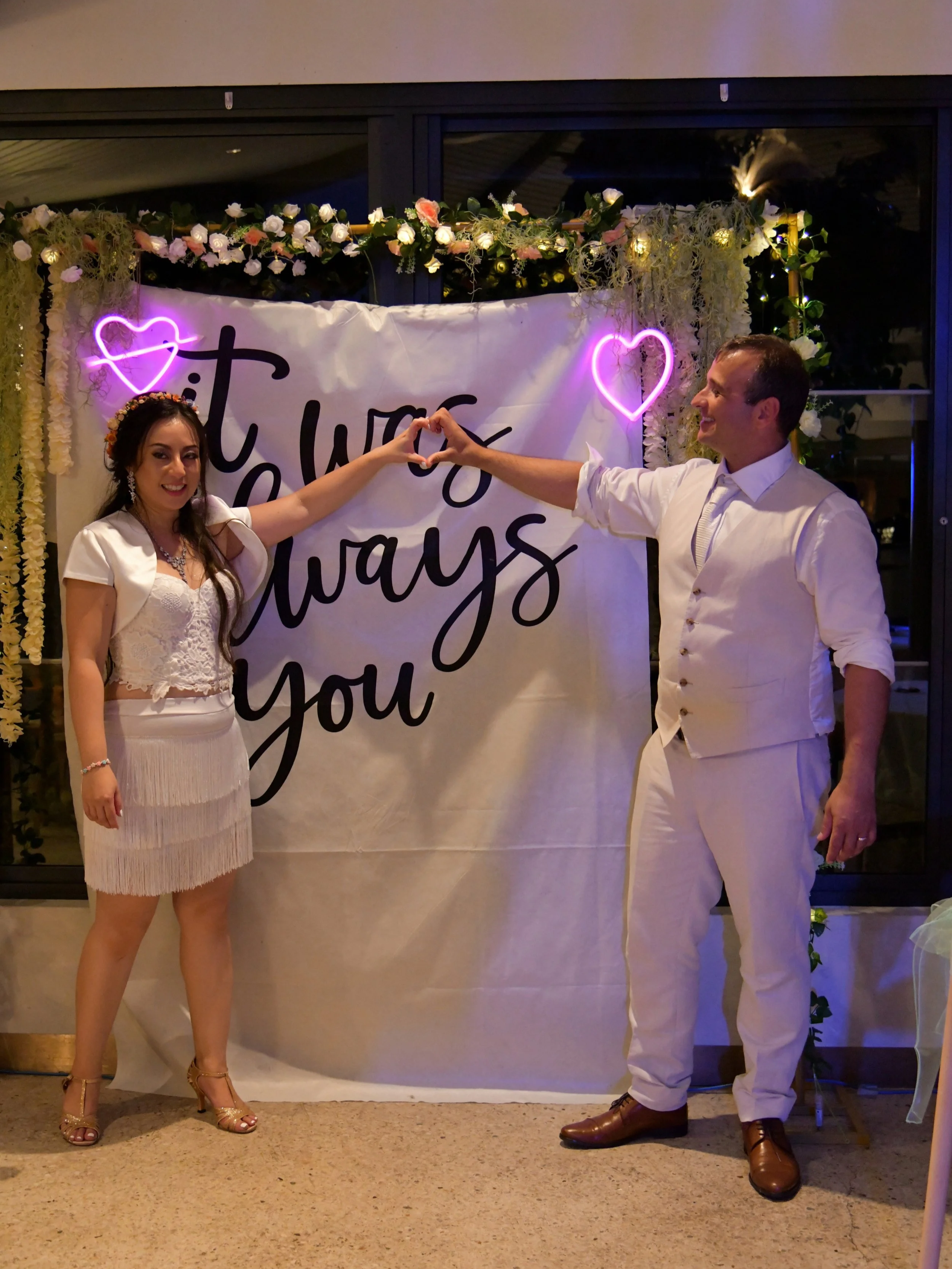 A couple, dressed in wedding attire, standing in front of a decorated backdrop with neon heart lights and the words "it was always you," holding hands and forming a heart with their arms.