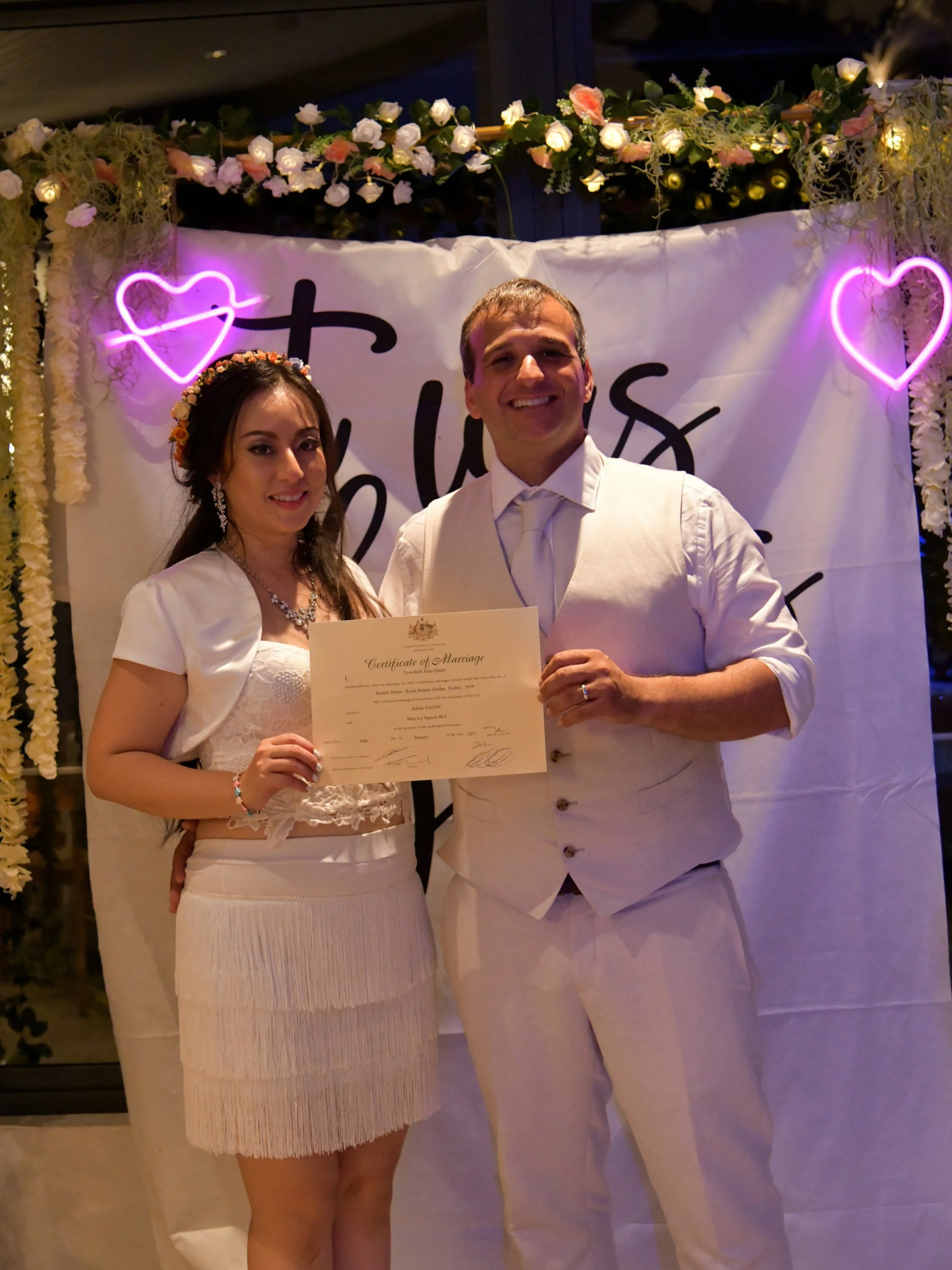 A couple dressed in white, holding a marriage certificate, stands in front of a decorated backdrop with purple neon hearts and flowers, celebrating their wedding.