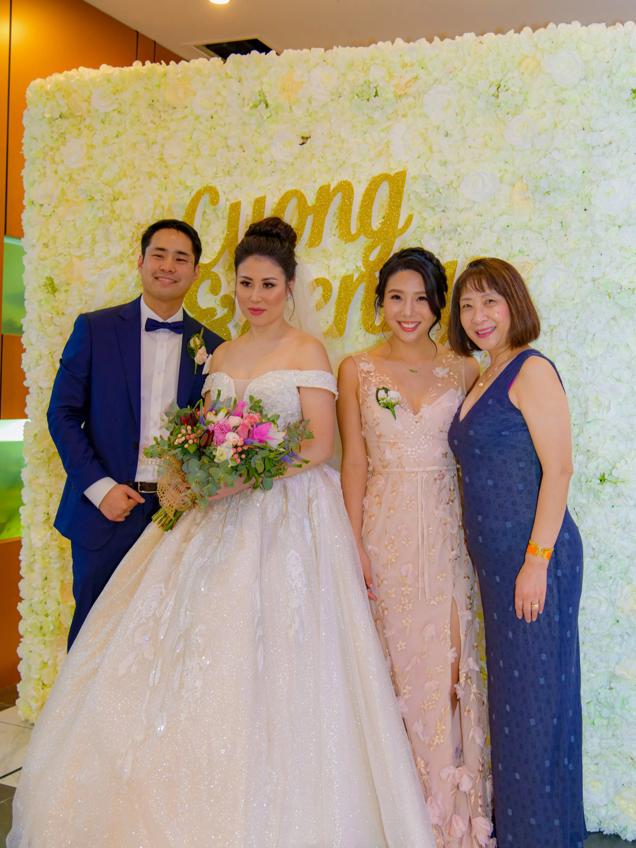 A group of four people at a wedding, standing in front of a white floral backdrop with the words 'Cung' and 'S' visible, smiling and posing for a photo. One woman is dressed in a white wedding gown holding a bouquet, and a man in a navy suit and bow 