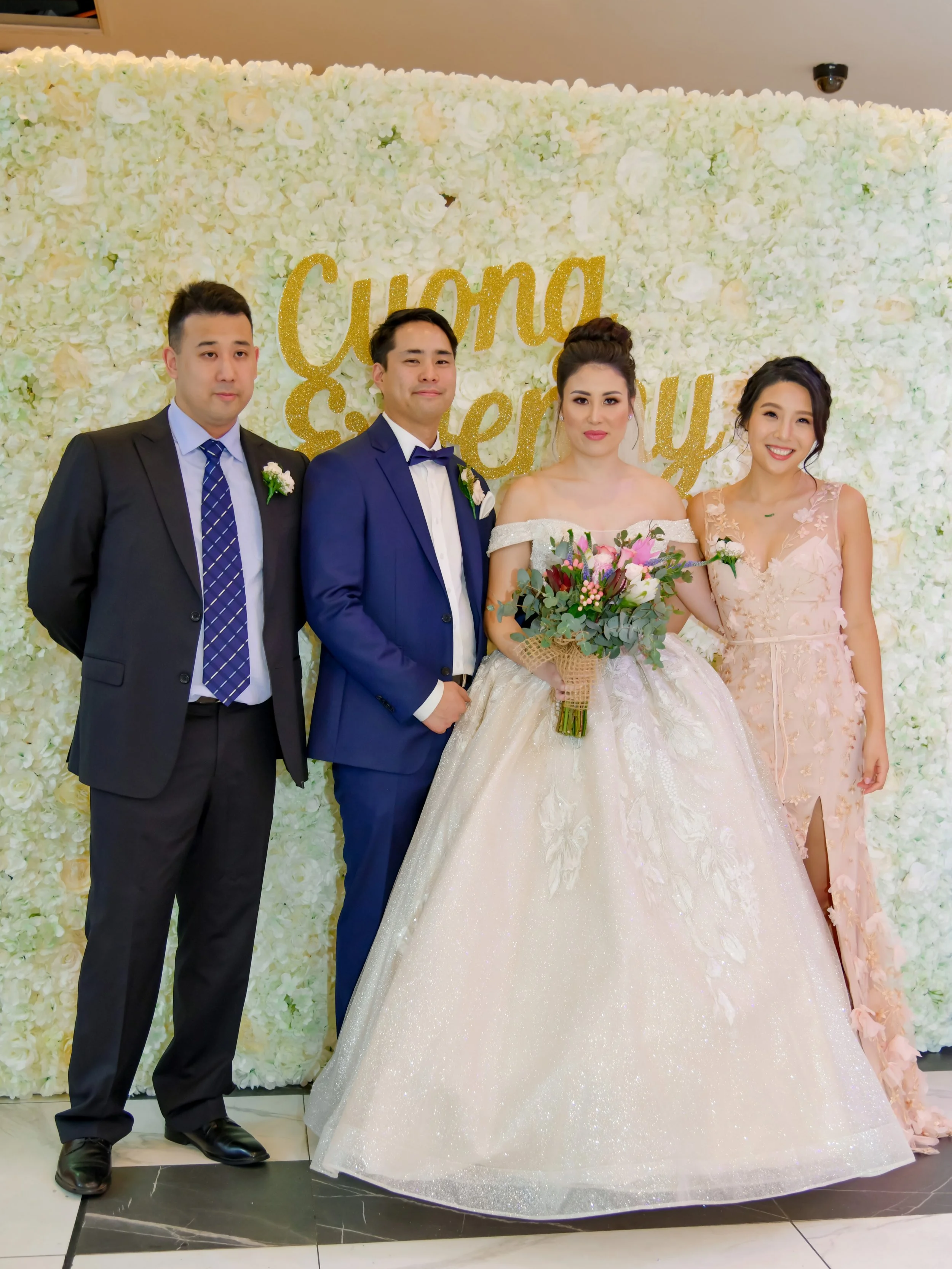 Four people standing in front of a wedding floral backdrop with gold text that reads 'Cuna and Sery' at a wedding celebration. The bride is in a ball gown holding a bouquet, the groom is in a blue suit, and two guests are dressed in formal suits and 