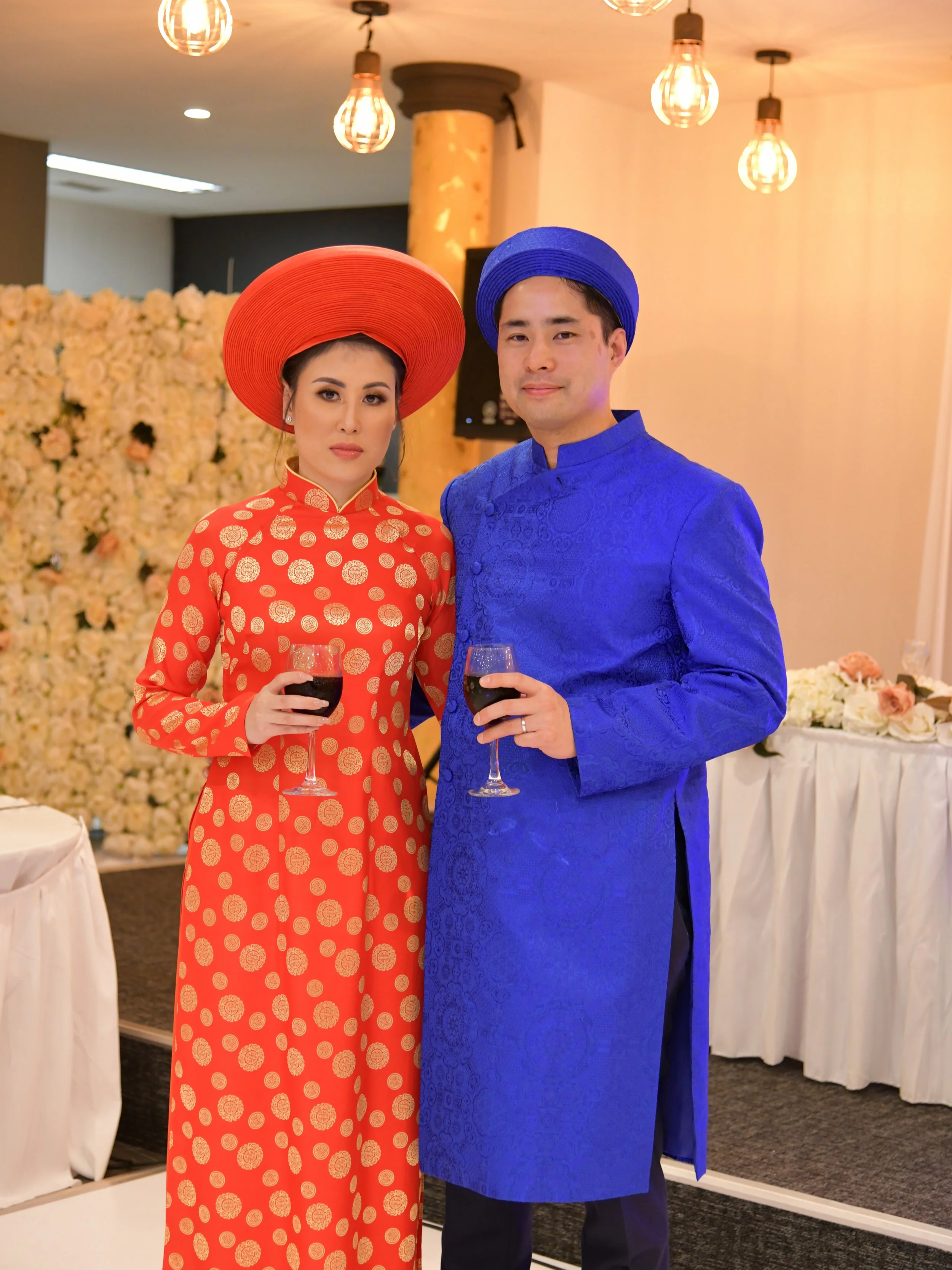 A couple dressed in traditional Vietnamese attire at a celebration, holding glasses of red wine, with a decorated floral backdrop and warm lighting.