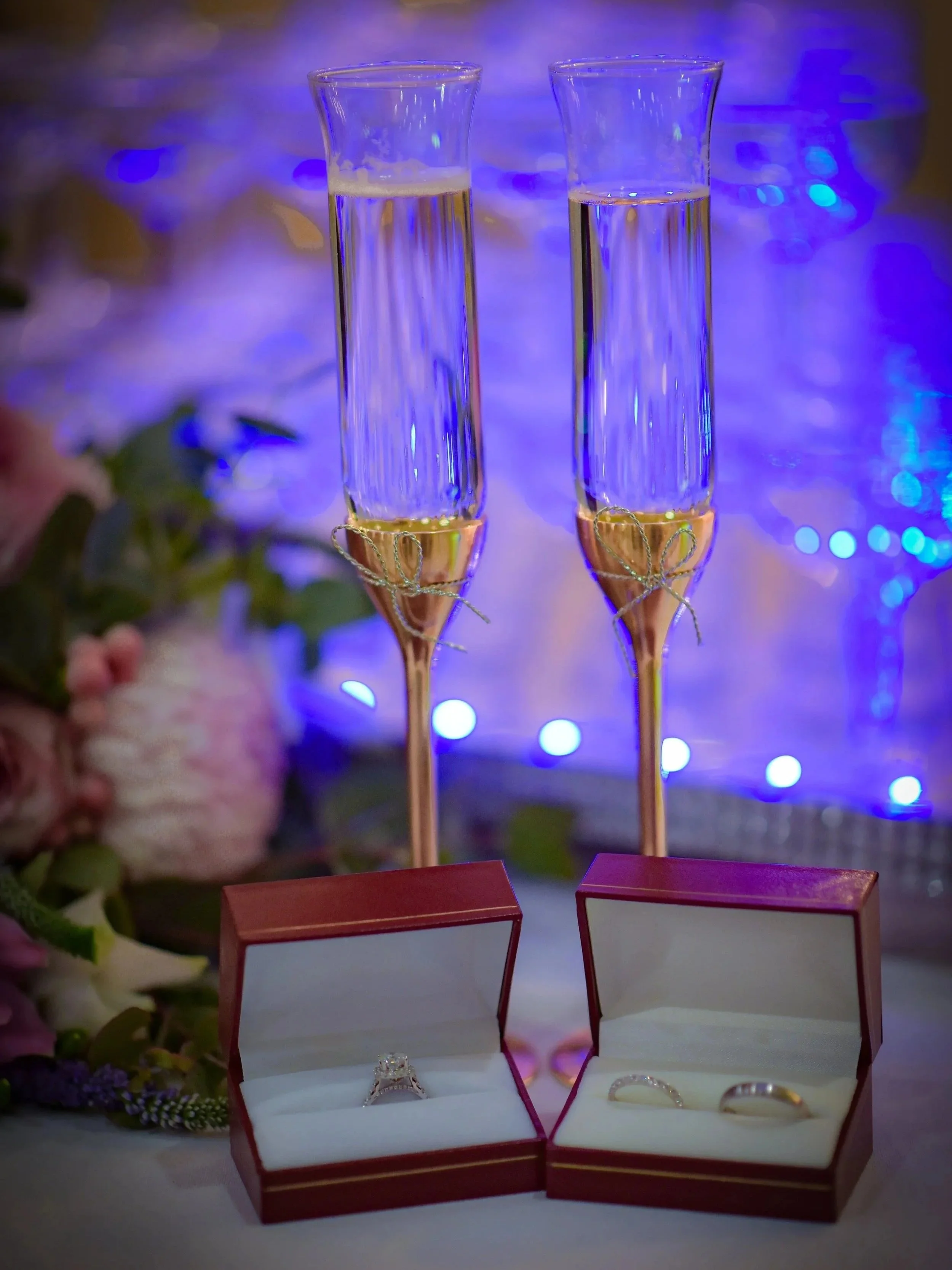 Two glasses of champagne decorated with gold ribbons and two wedding rings in open red boxes on a table with blurred flowers and blue lights in the background.