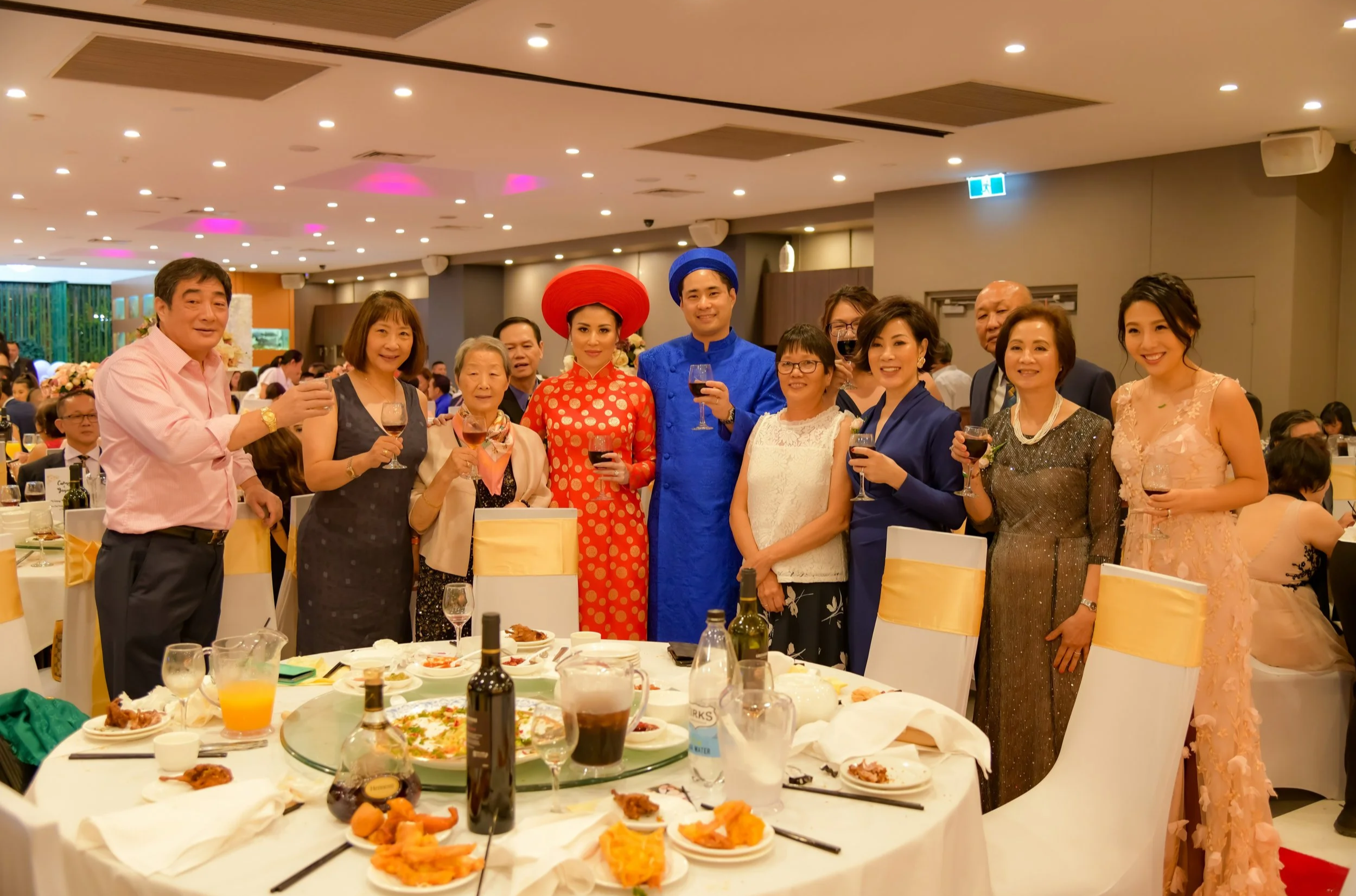 Group of people celebrating at a banquet, some in traditional Vietnamese clothing and accessories, holding wine glasses and smiling.