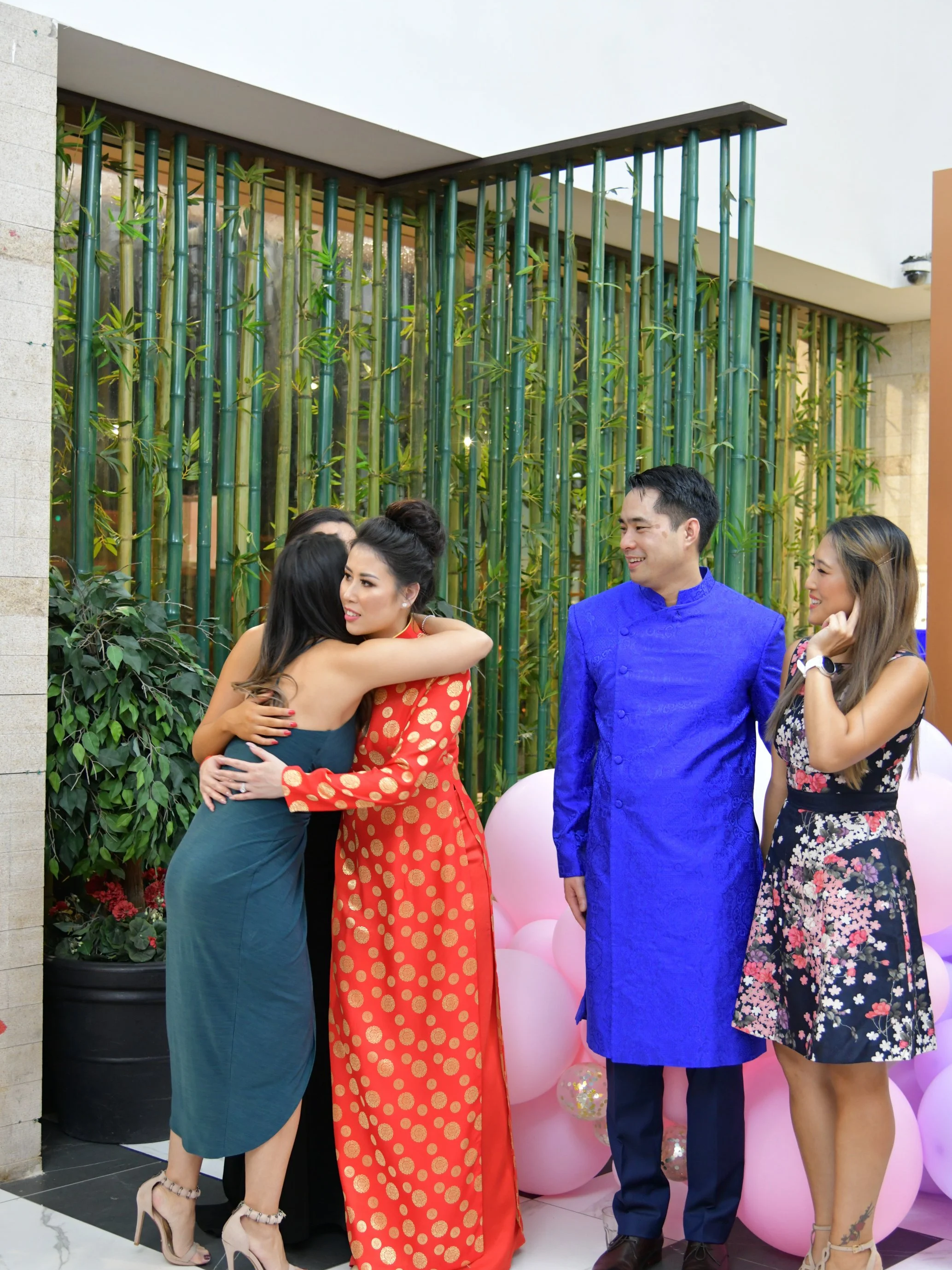 People hugging and smiling at a celebration with pink balloons and bamboo decorations.