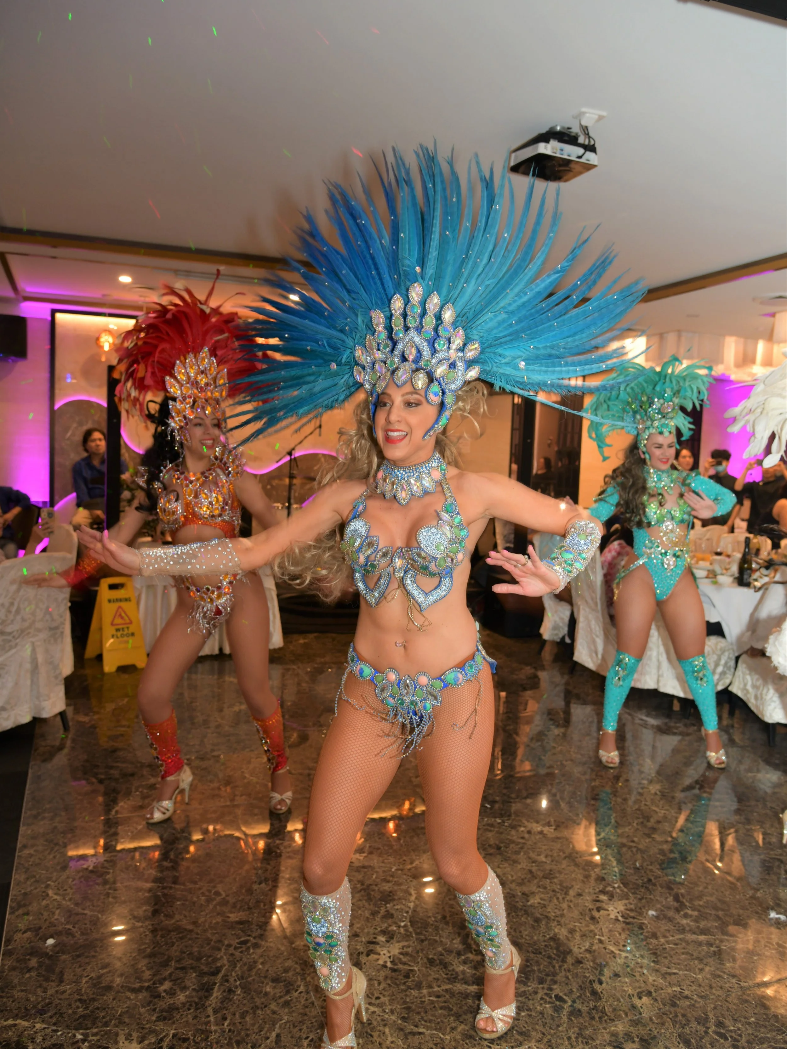 Women performing a dance in colorful, elaborate carnival costumes with large feathered headdresses and sparkling accessories inside a decorated venue.