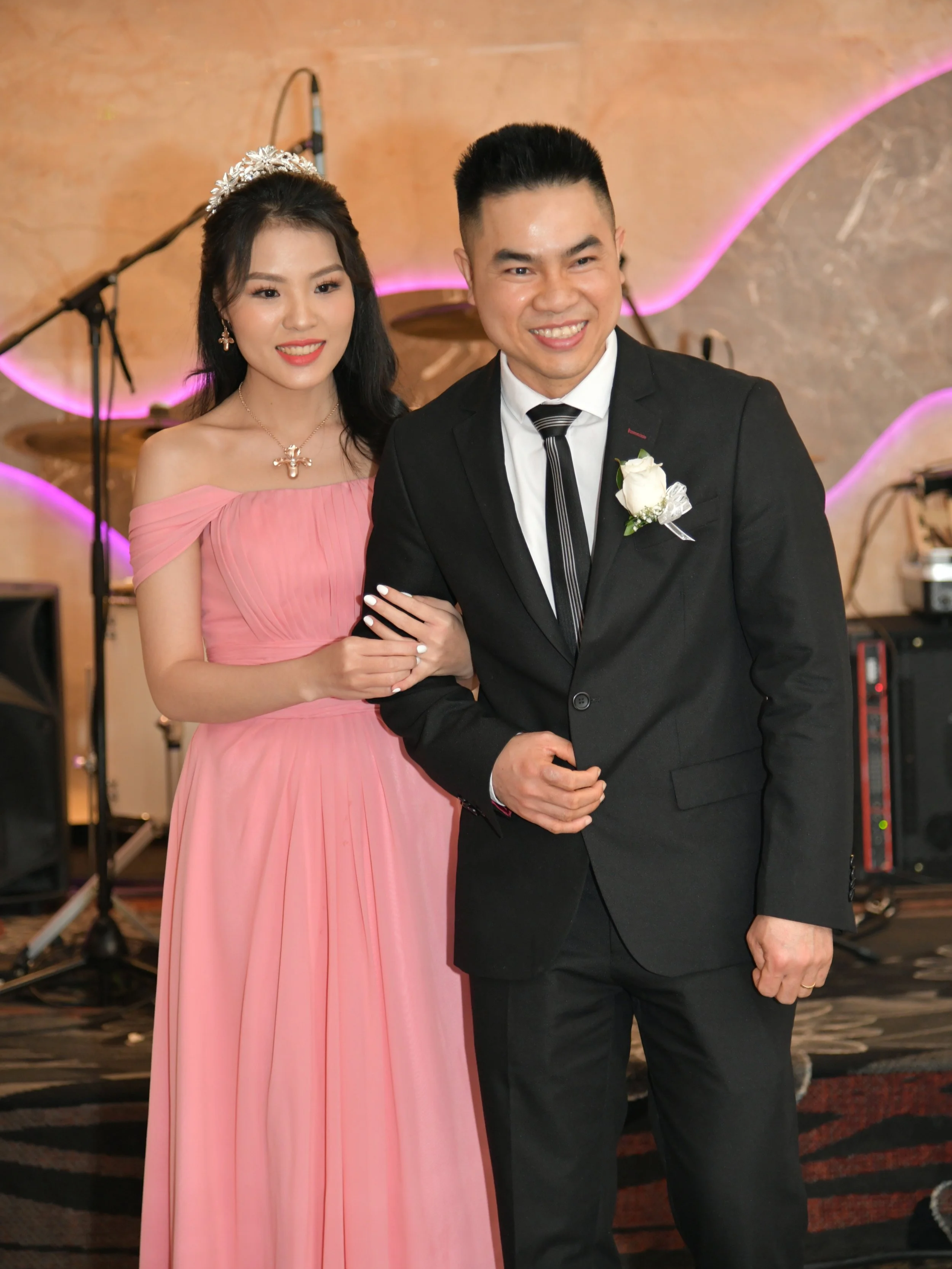 A couple dressed in formal attire, the woman in a pink gown with a tiara and jewelry, and the man in a black suit with a white shirt, striped tie, and boutonniere, smiling together at an event with musical instruments in the background.