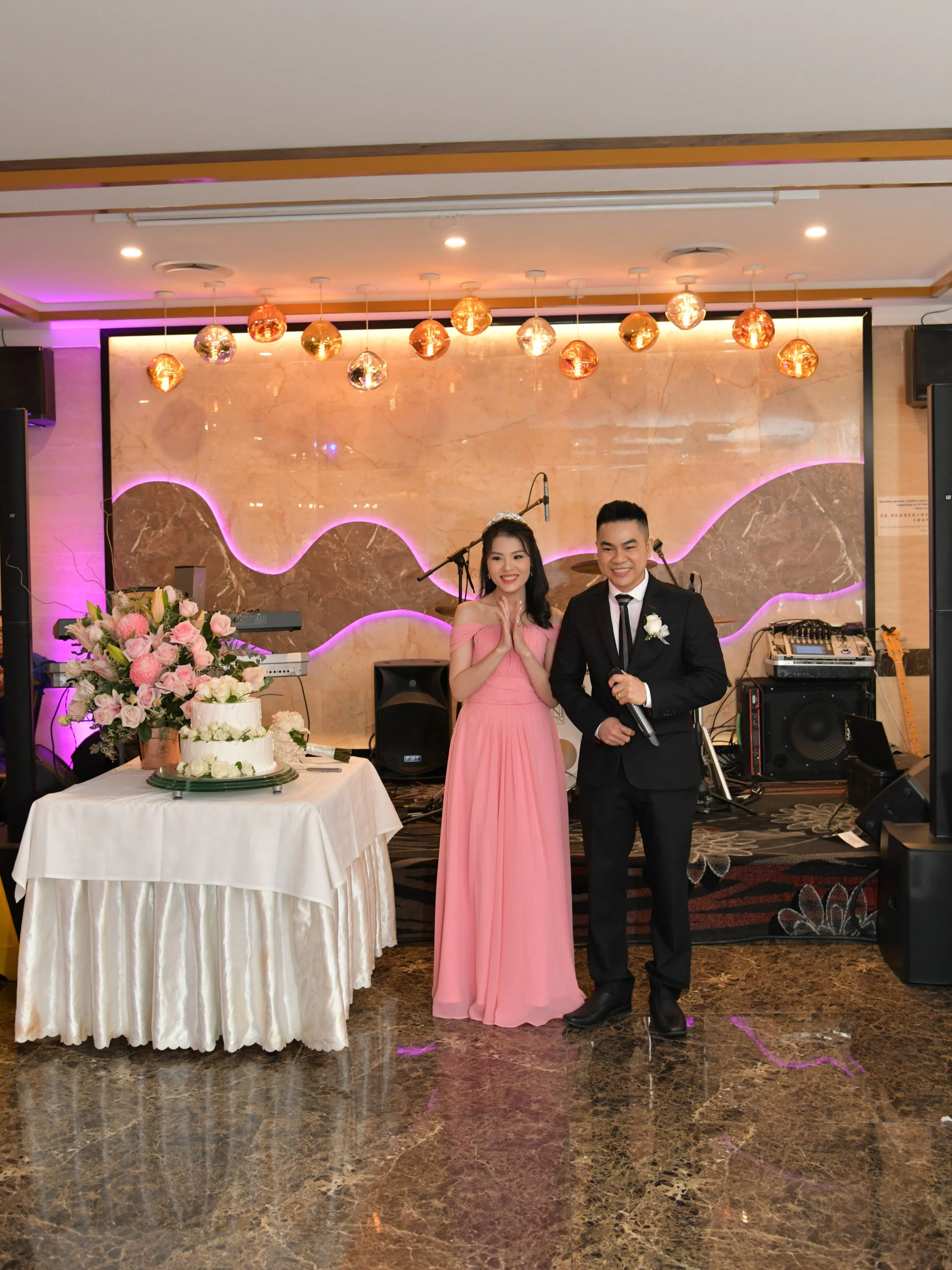 A couple dressed in formal attire standing in front of a decorated stage, with a table of flowers and cake on the left, during a celebration or wedding.