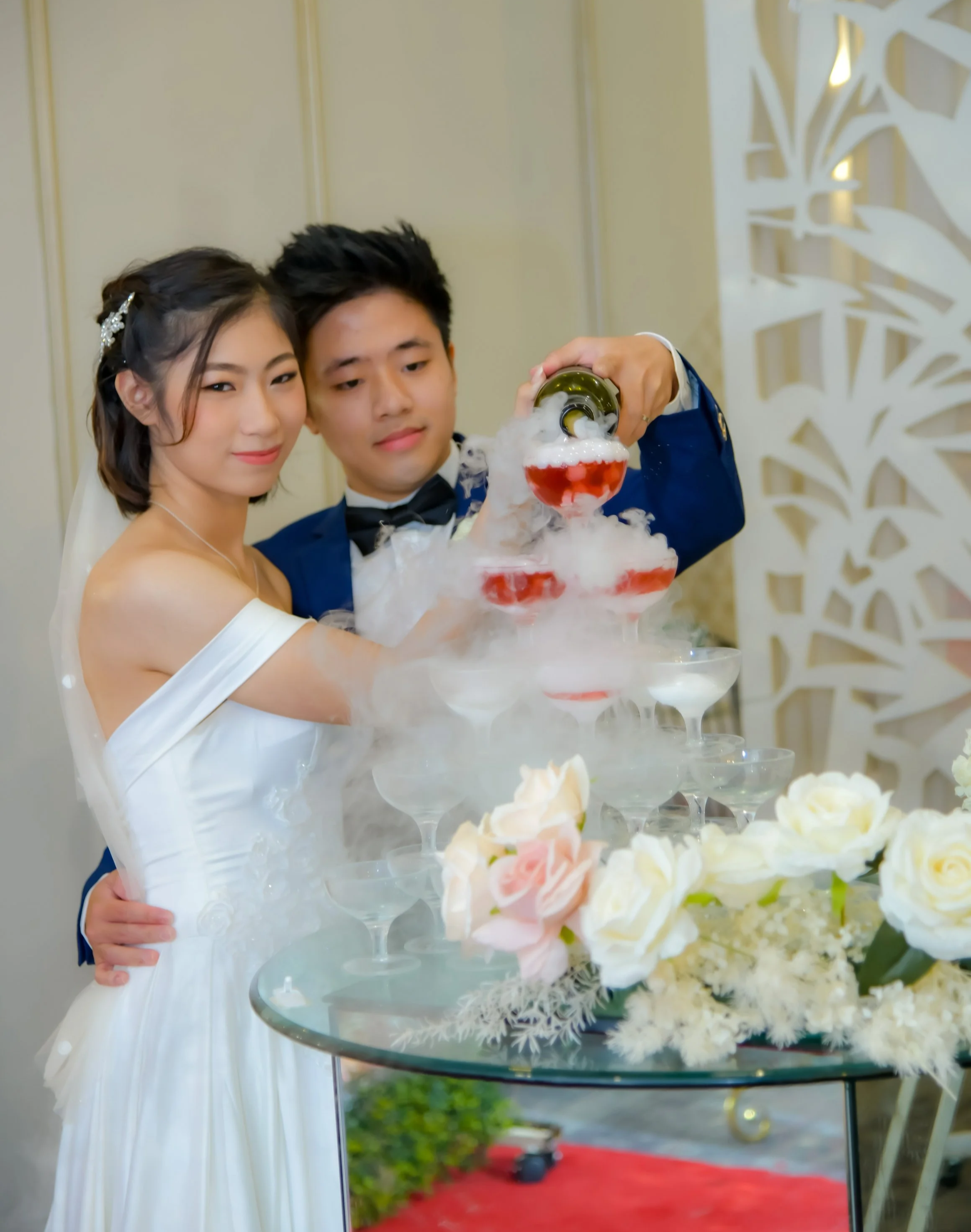 Bride and groom in wedding attire pouring champagne into a stacked pyramid of glasses with dry ice effects, white and pink roses on a glass table, elegant wedding setting.