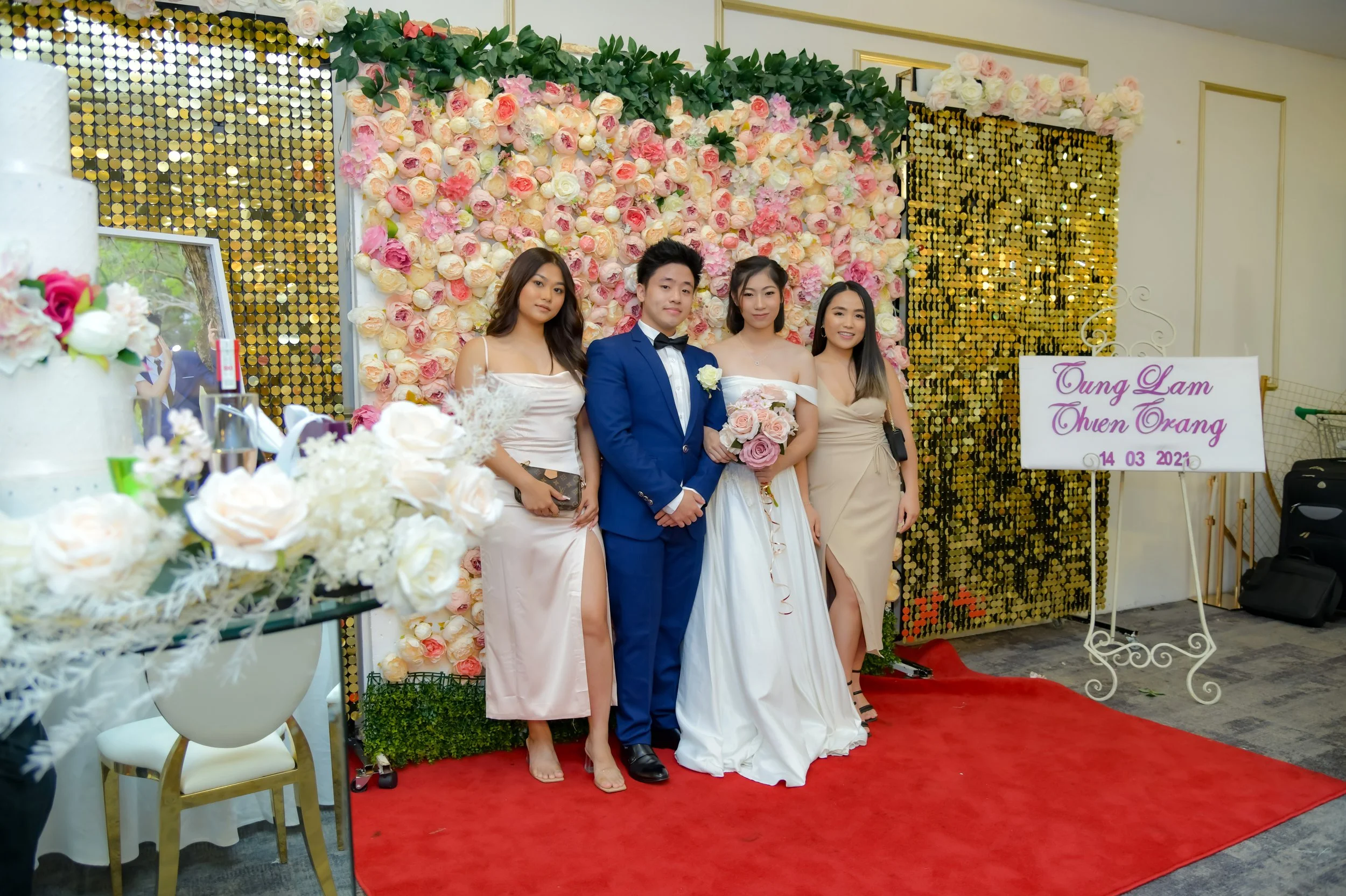 Group of four people, two women in dresses and a man in a tuxedo, standing on a red carpet in front of a floral backdrop at a wedding reception. The woman in the center is holding a bouquet of pink and white roses. There is a sign with purple text th