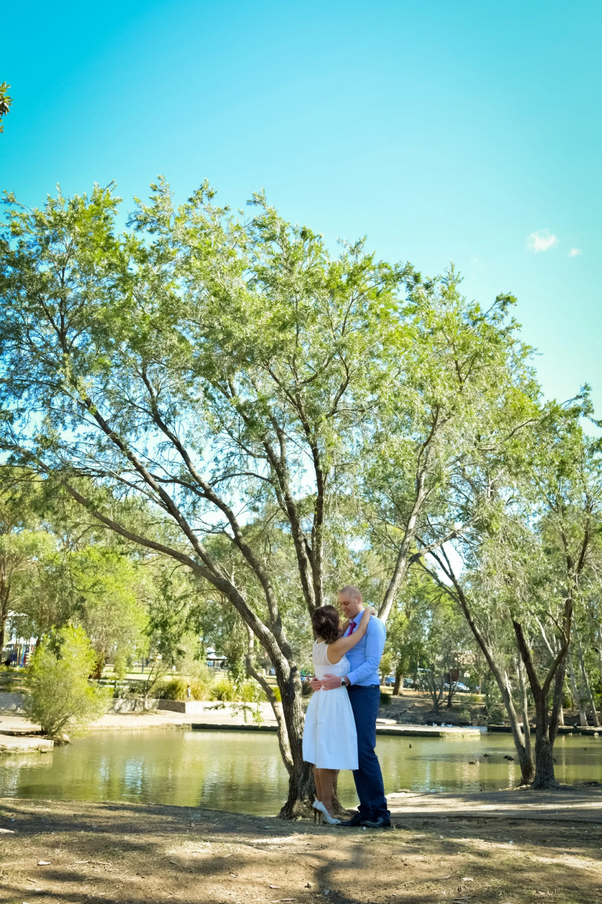 A couple in wedding attire standing close and embracing near a tree by a pond in a park.