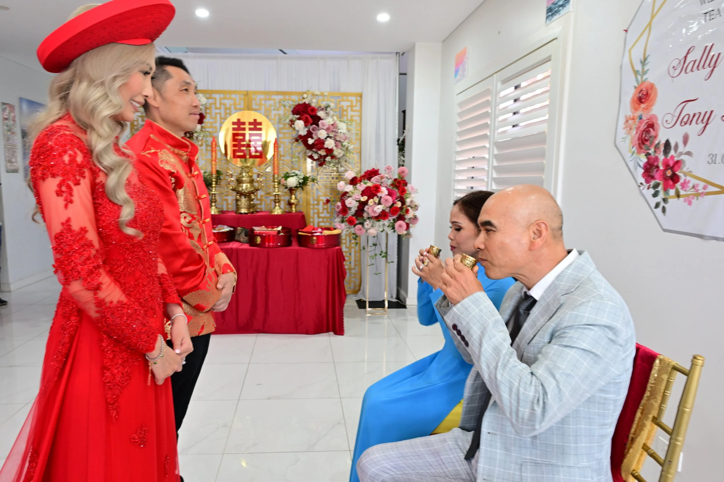 Two women and one man in traditional red Chinese wedding attire standing and facing two seated people, a man in a gray suit and a woman in a blue dress, during a wedding ceremony with flowers and celebratory decorations in the background.