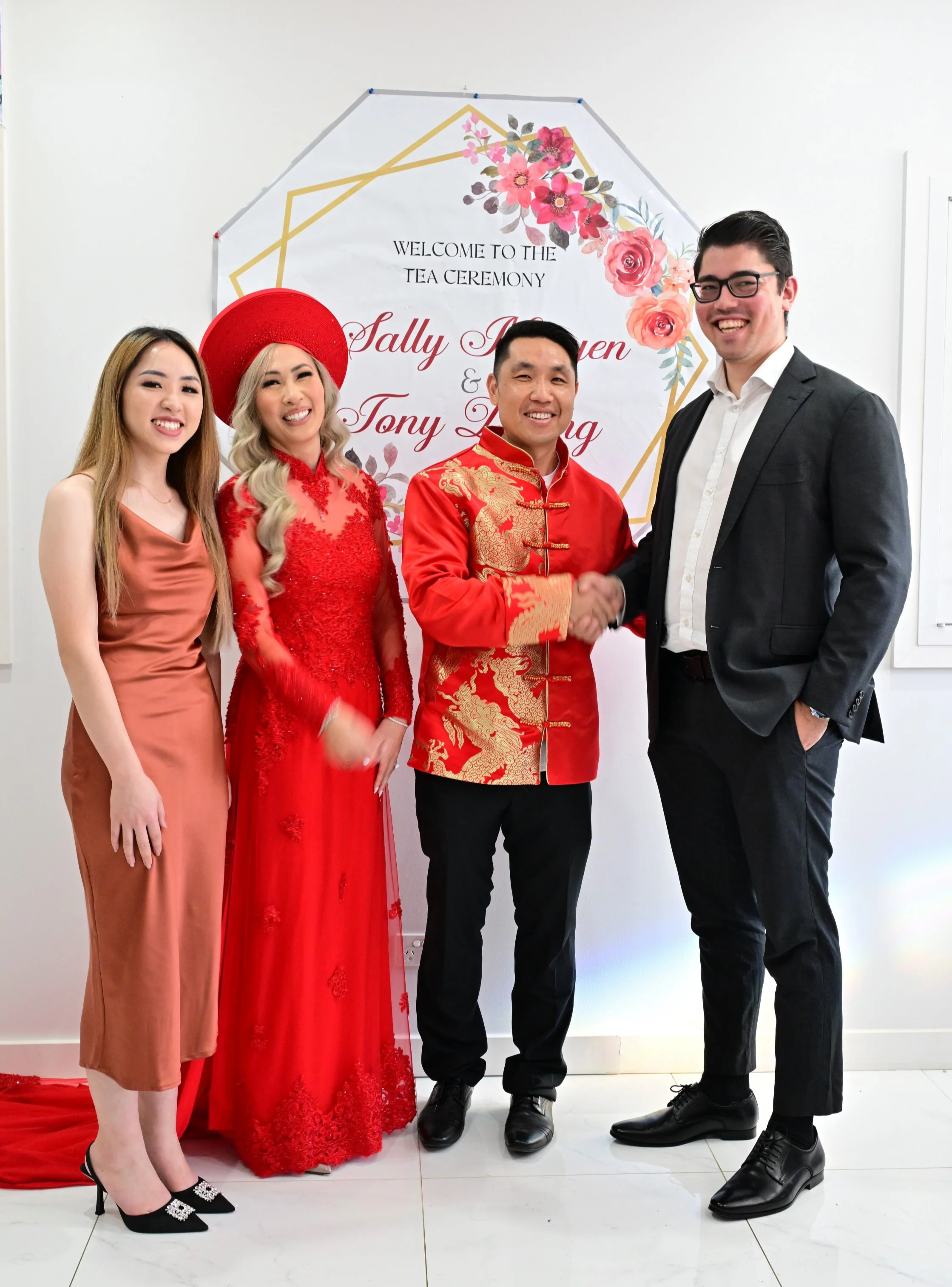 A group of four people, two women and two men, standing in front of a wedding sign, with the man on the right and the woman in the red traditional dress shaking hands. The wedding sign reads, 'Welcome to the tea ceremony, Sally Nguyen & Tony Leong,' 