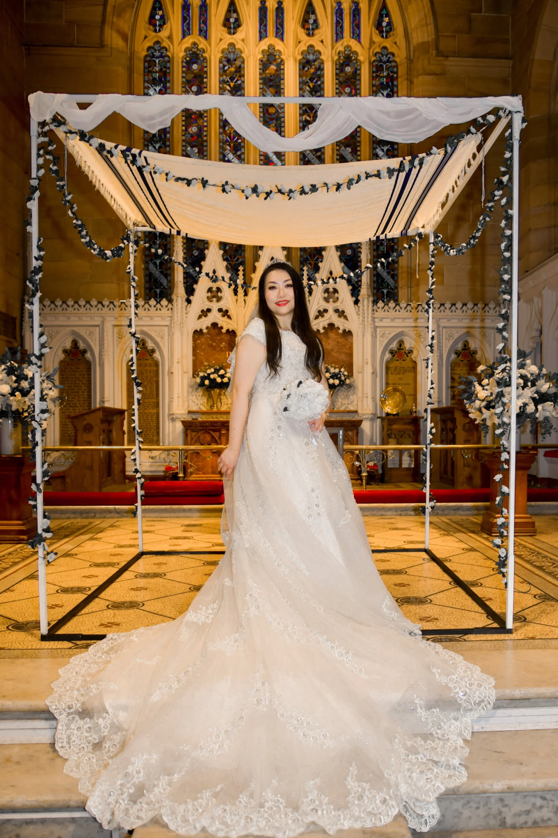 A bride in a white wedding gown holding a bouquet, standing inside a decorated church with stained glass windows and ornate woodwork.