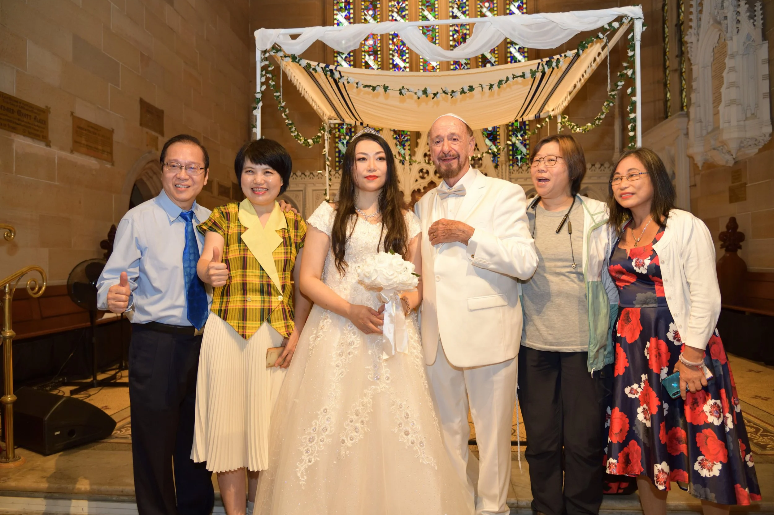 Group of six people, including a bride in a white wedding dress holding a bouquet and an older man in a white suit, standing together inside a church. The group appears to be celebrating, with some giving thumbs up. The church has stained glass windo