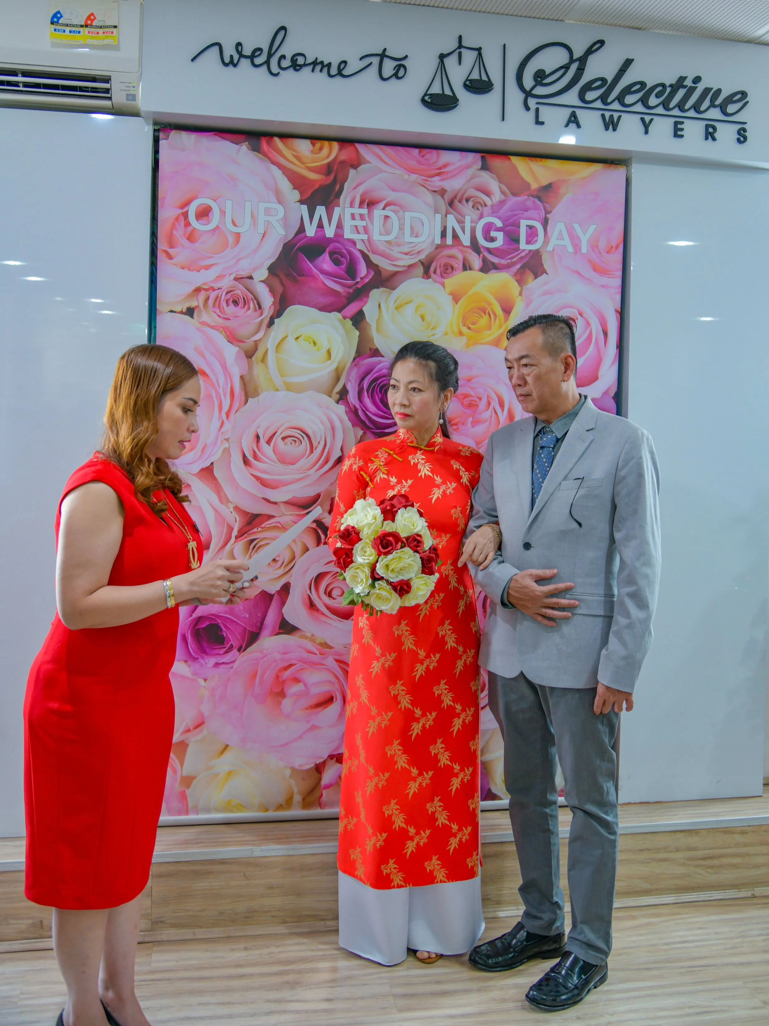 A couple is getting married inside a wedding chapel, with a woman in a red ao dai (traditional Vietnamese dress) holding a bouquet of flowers. They are signing a wedding document, standing in front of a floral backdrop that reads 'Our Wedding Day,' w