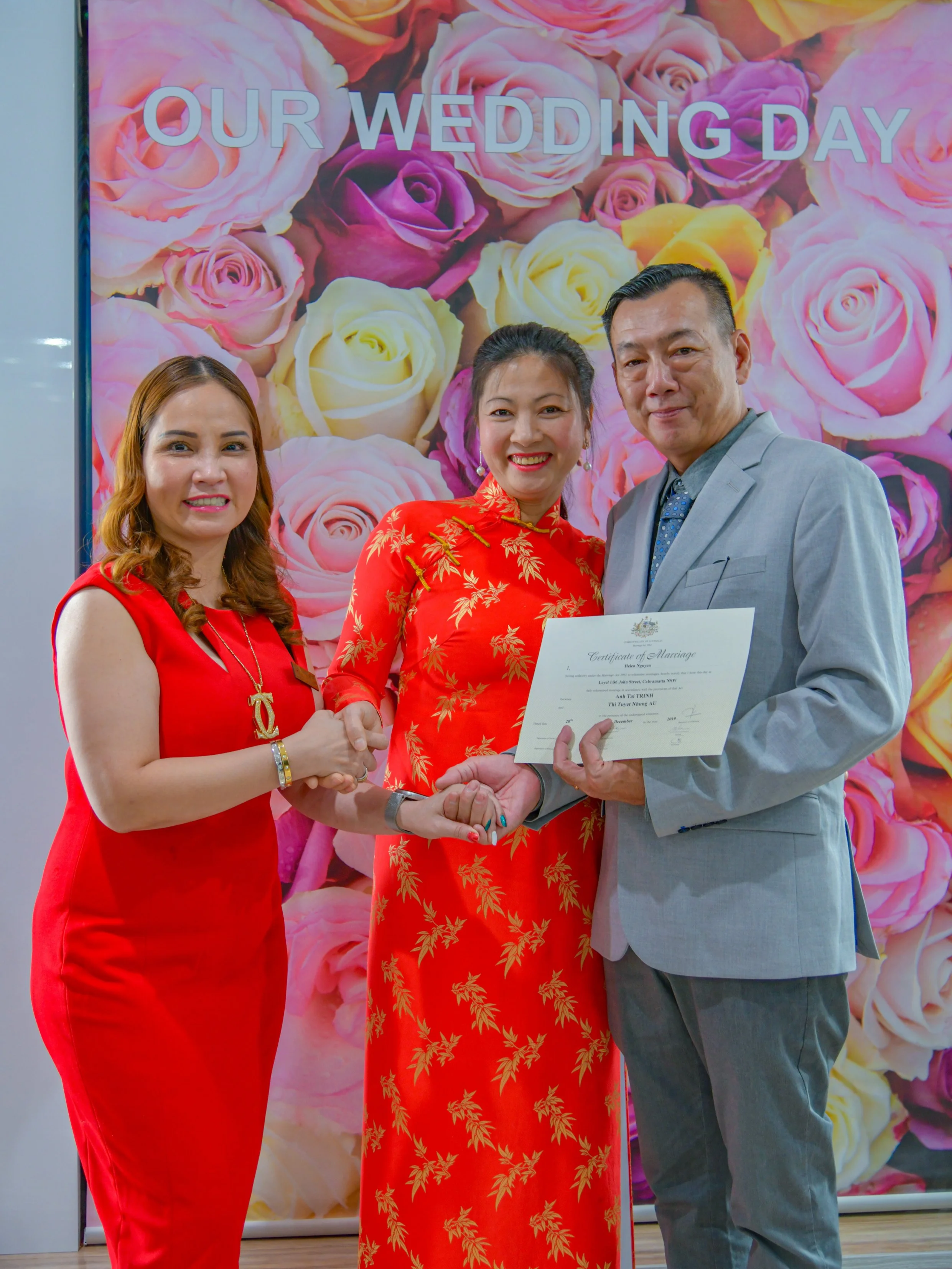 A wedding ceremony with a couple holding a marriage certificate, flanked by a woman in a red dress, in front of a backdrop with roses and the text "Our Wedding Day."