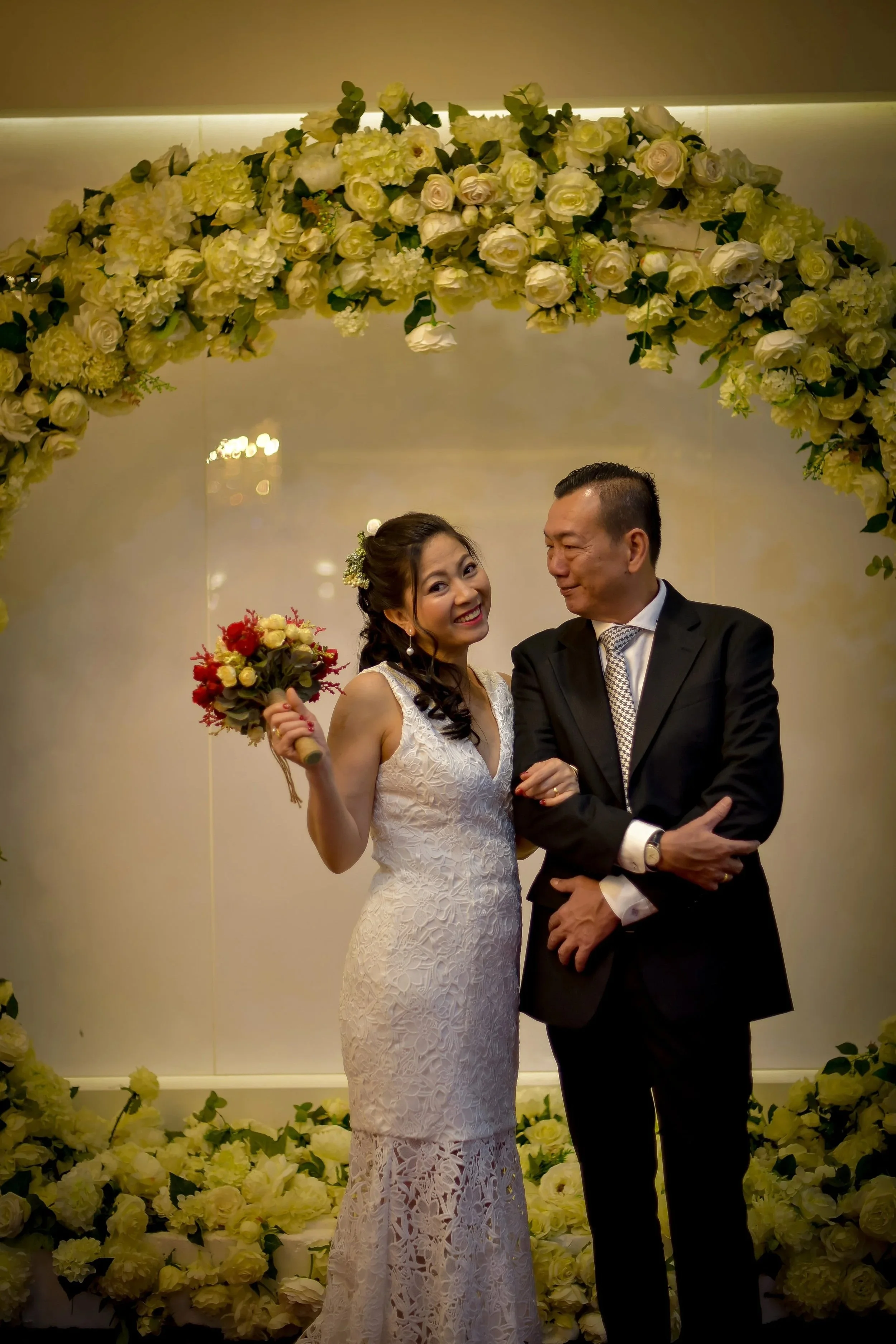 A smiling woman in a white lace wedding dress holding a bouquet of flowers, standing next to a man in a black suit underneath a flower arch at a wedding ceremony.