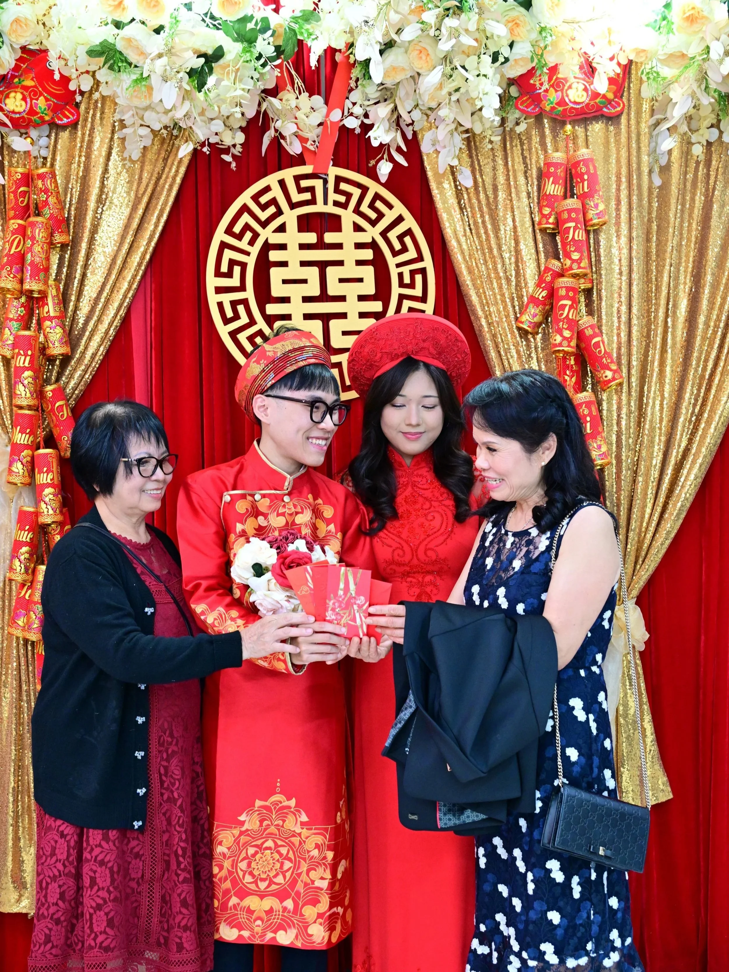 A traditional Vietnamese wedding ceremony with the bride and groom dressed in red, receiving red envelopes and flowers from family members in front of a red and gold decorated backdrop with flowers, curtains, and a double happiness symbol.