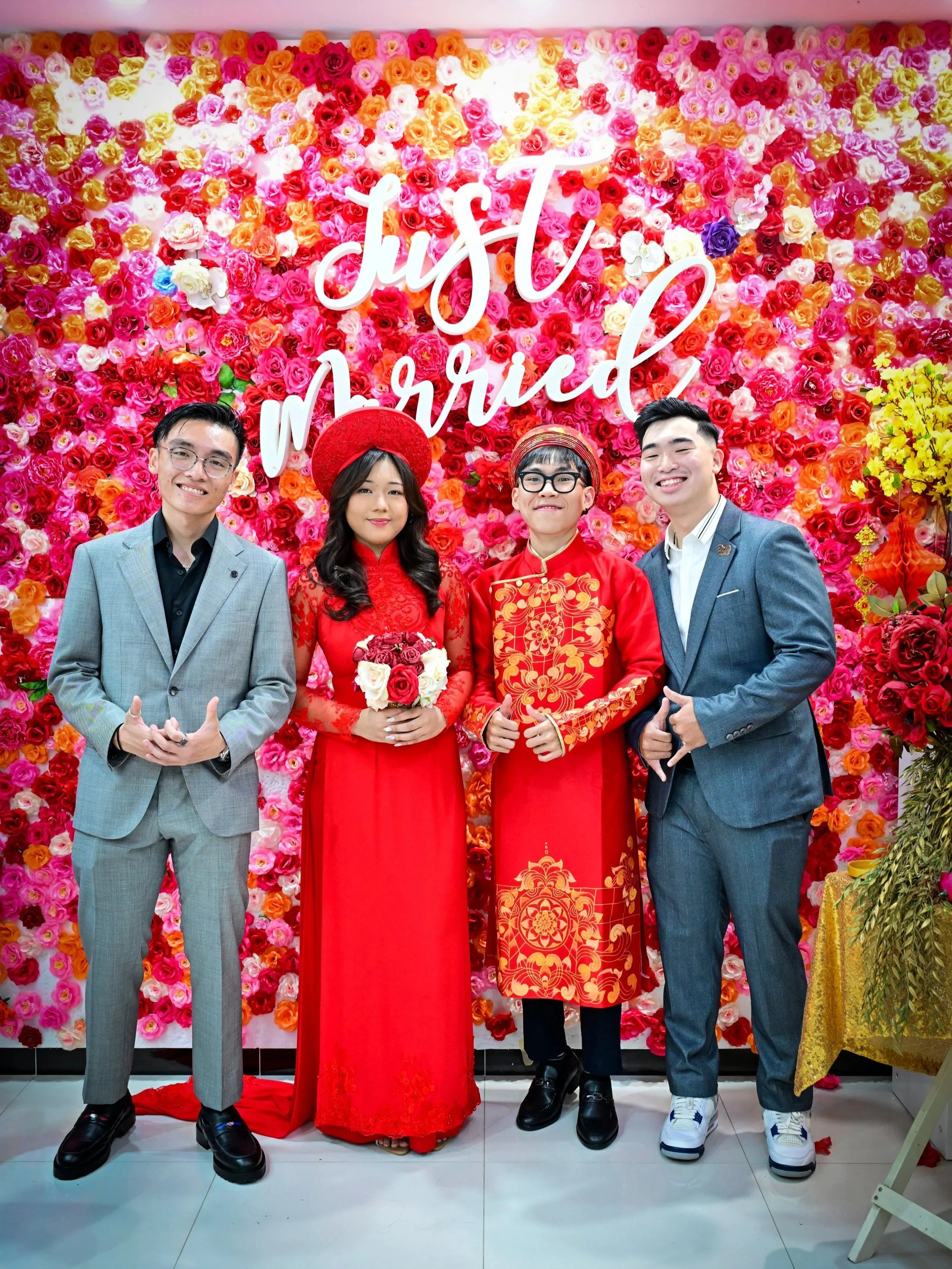 Four people standing in front of a colorful floral wall with a neon sign that reads 'Just Married.' The couple in the center is dressed in traditional Vietnamese wedding attire, with the woman in a red Ao Dai and the man in a red embroidered tunic. T
