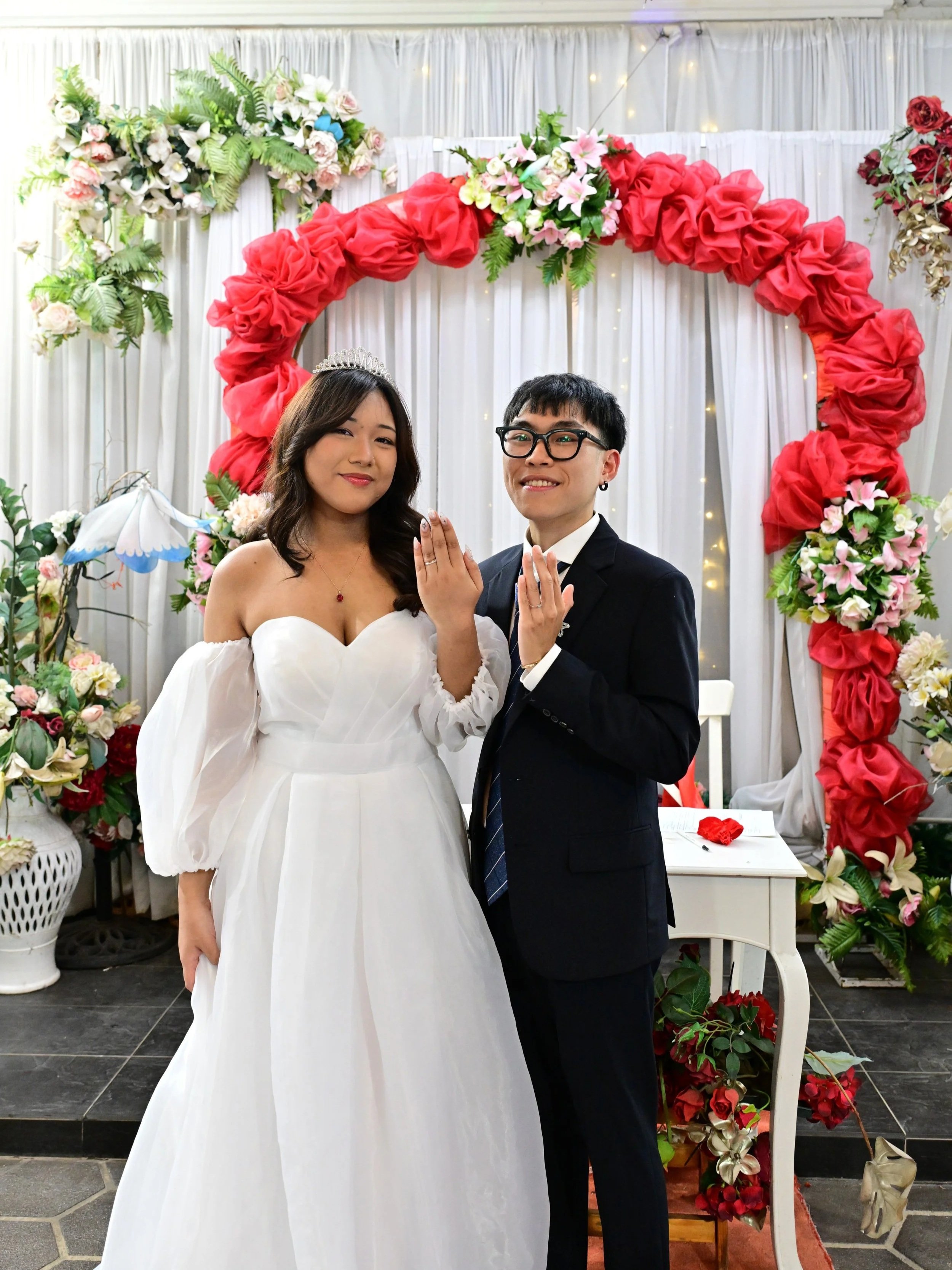 A couple exchanging wedding rings during their wedding ceremony, standing in front of a floral wedding arch decorated with pink, red, and white flowers.