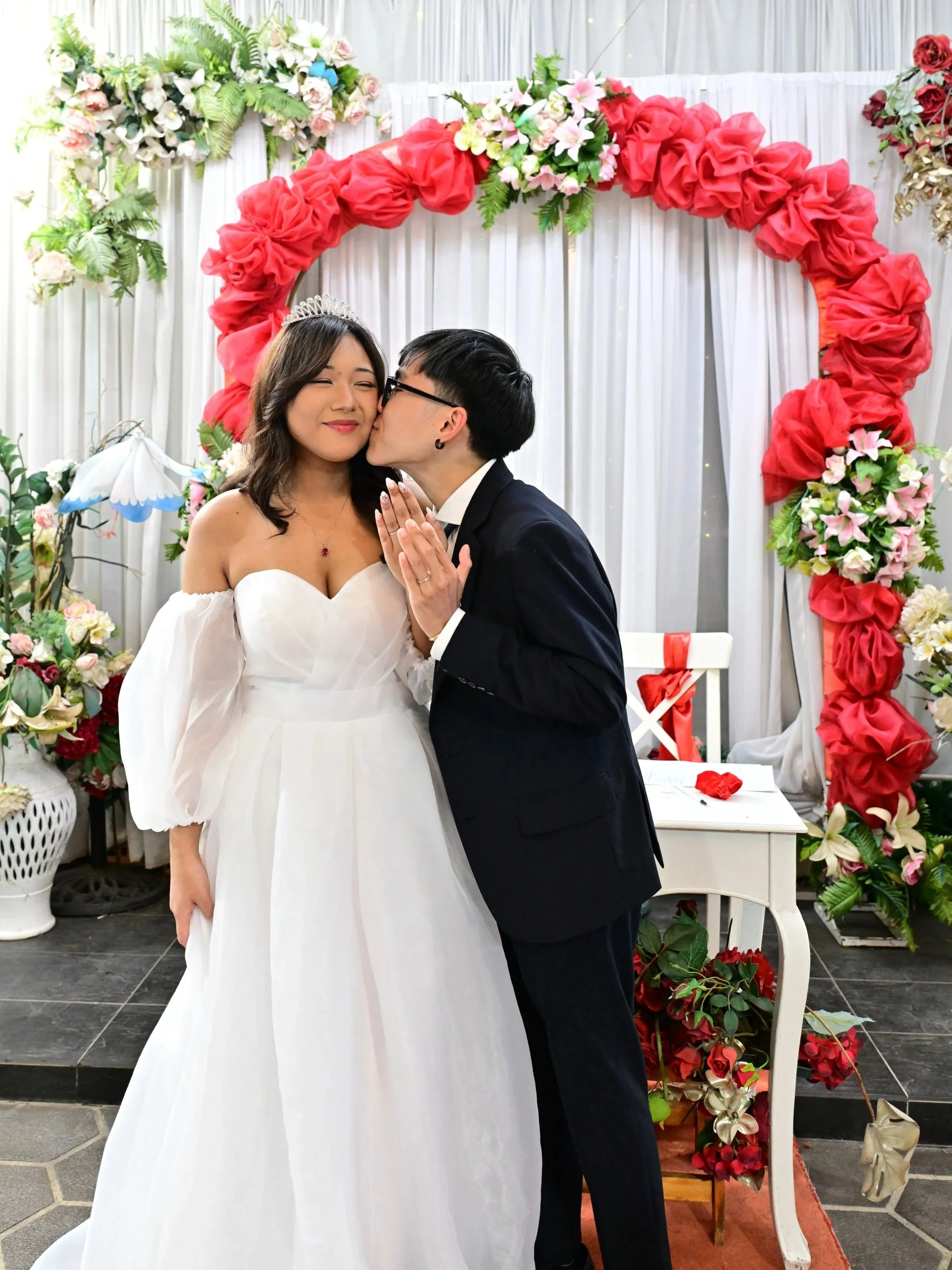 A bride in a white wedding gown and a groom in a black suit sharing a moment at their wedding, with a floral heart-shaped backdrop decorated with pink and white flowers.