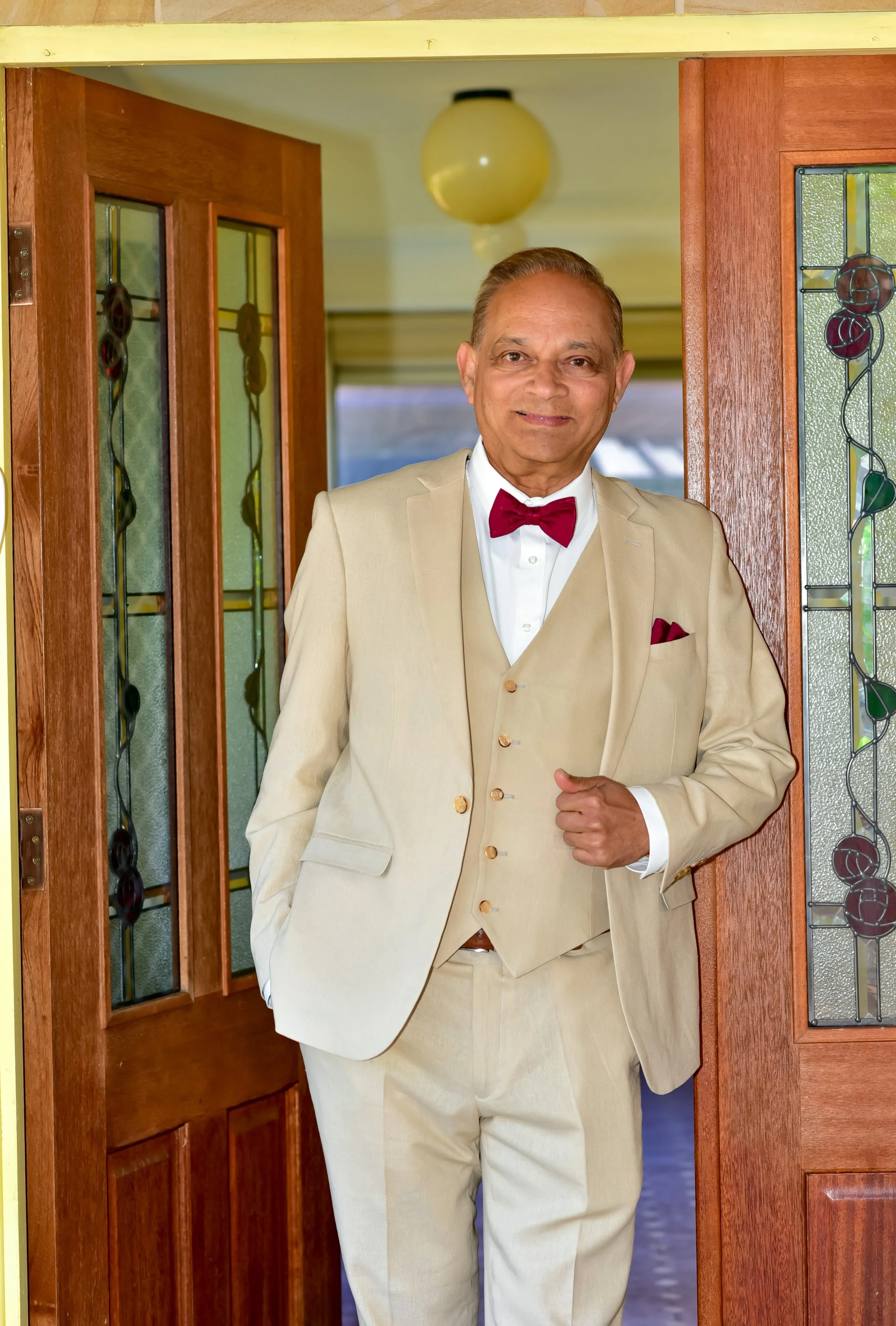 An elderly man dressed in a beige suit with a matching vest, white shirt, and red bow tie stands at a doorway with stained glass panels. He has his right hand in his pocket and is smiling.