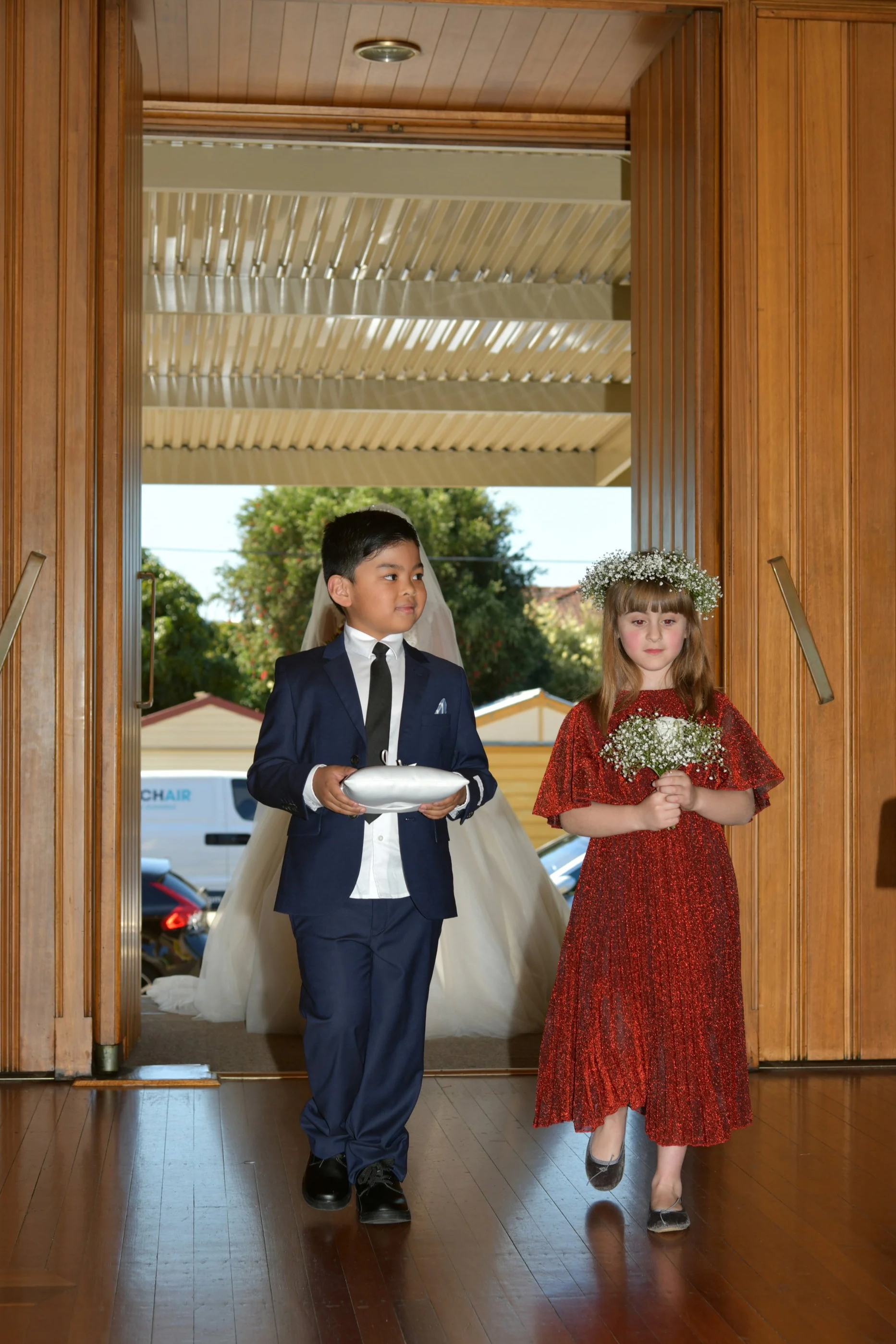 Children participating in a wedding ceremony, with a boy in a navy suit holding a silver pillow and a girl in a red dress holding a bouquet, walking through a wooden doorway.