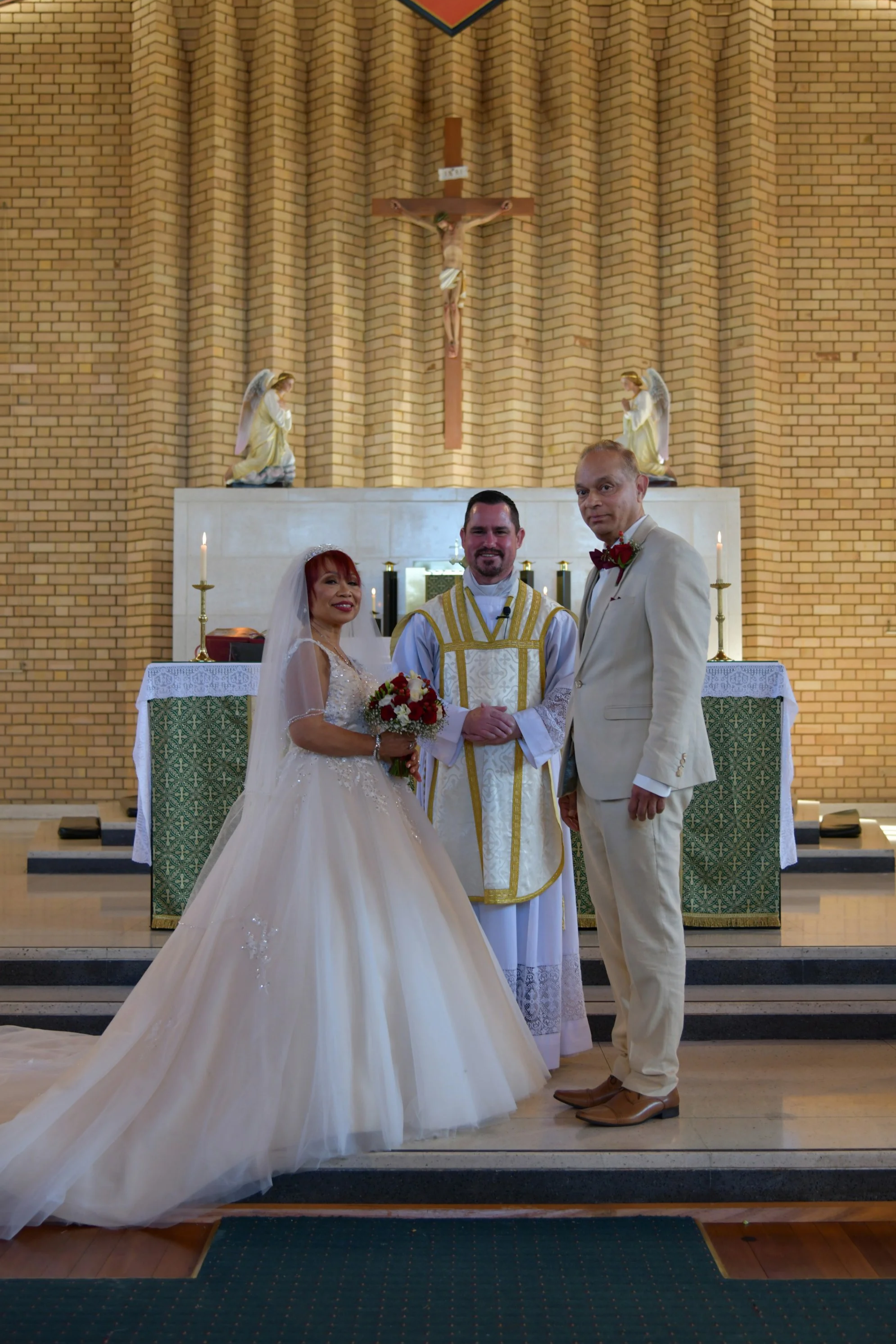 A bride and groom standing with a priest inside a church altar. The bride wears a white wedding gown and veil, holding a bouquet. The groom is dressed in a light-colored suit with a red bow tie. The priest is dressed in white and gold liturgical vest
