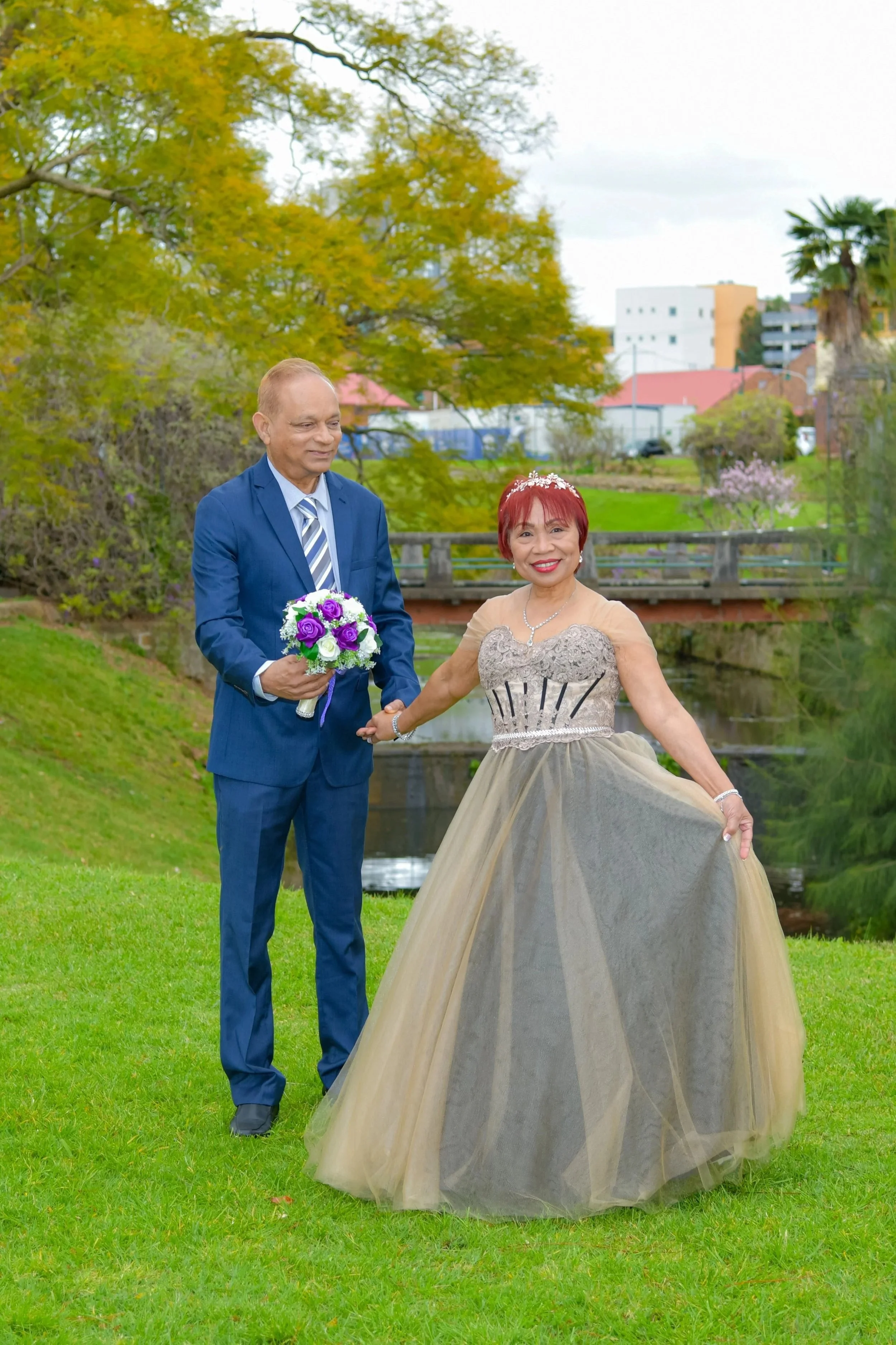 A woman in an elegant gown and a man in a blue suit holding a bouquet of purple and white flowers outside in a park-like setting with trees, grass, and a small bridge.