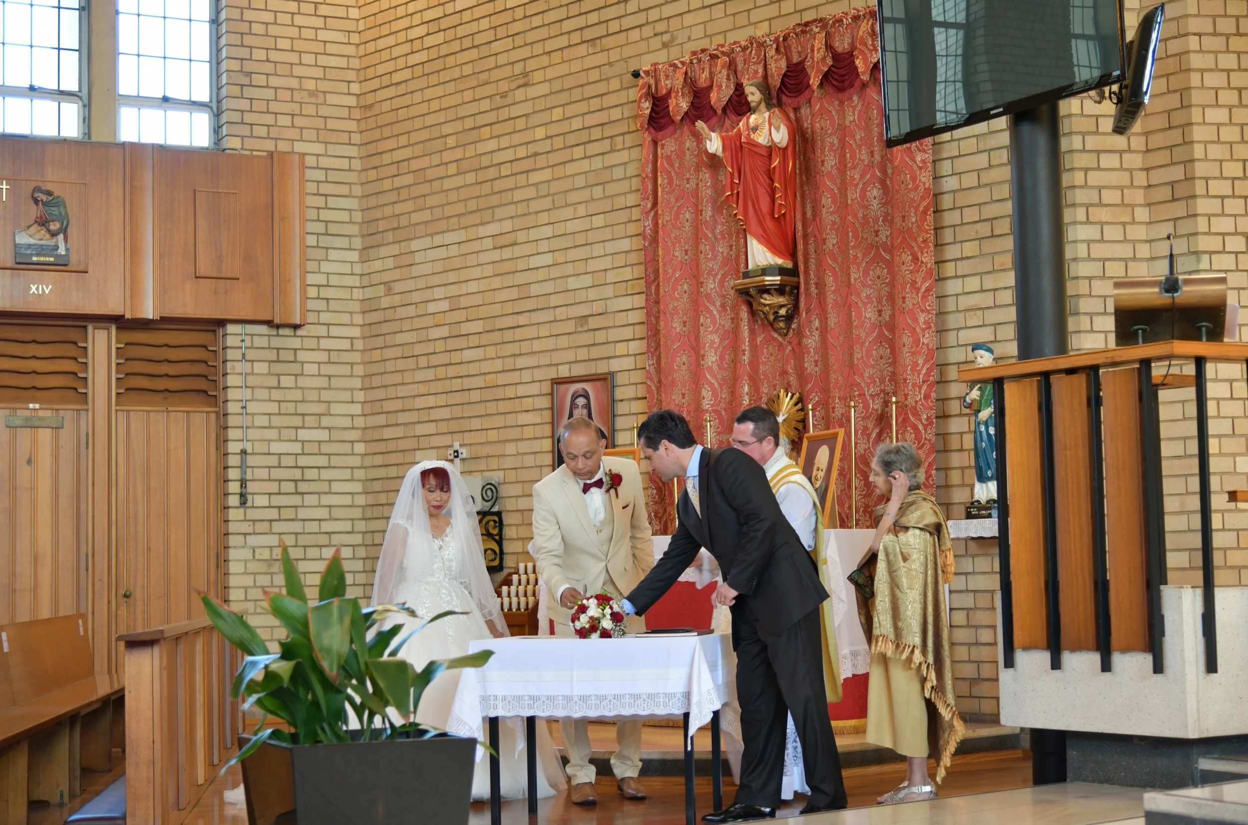 A wedding ceremony taking place inside a church with a brick wall background. The bride is dressed in a white wedding gown with a veil, and the groom is in a black suit. They are signing a marriage certificate at a table decorated with a flower bouqu
