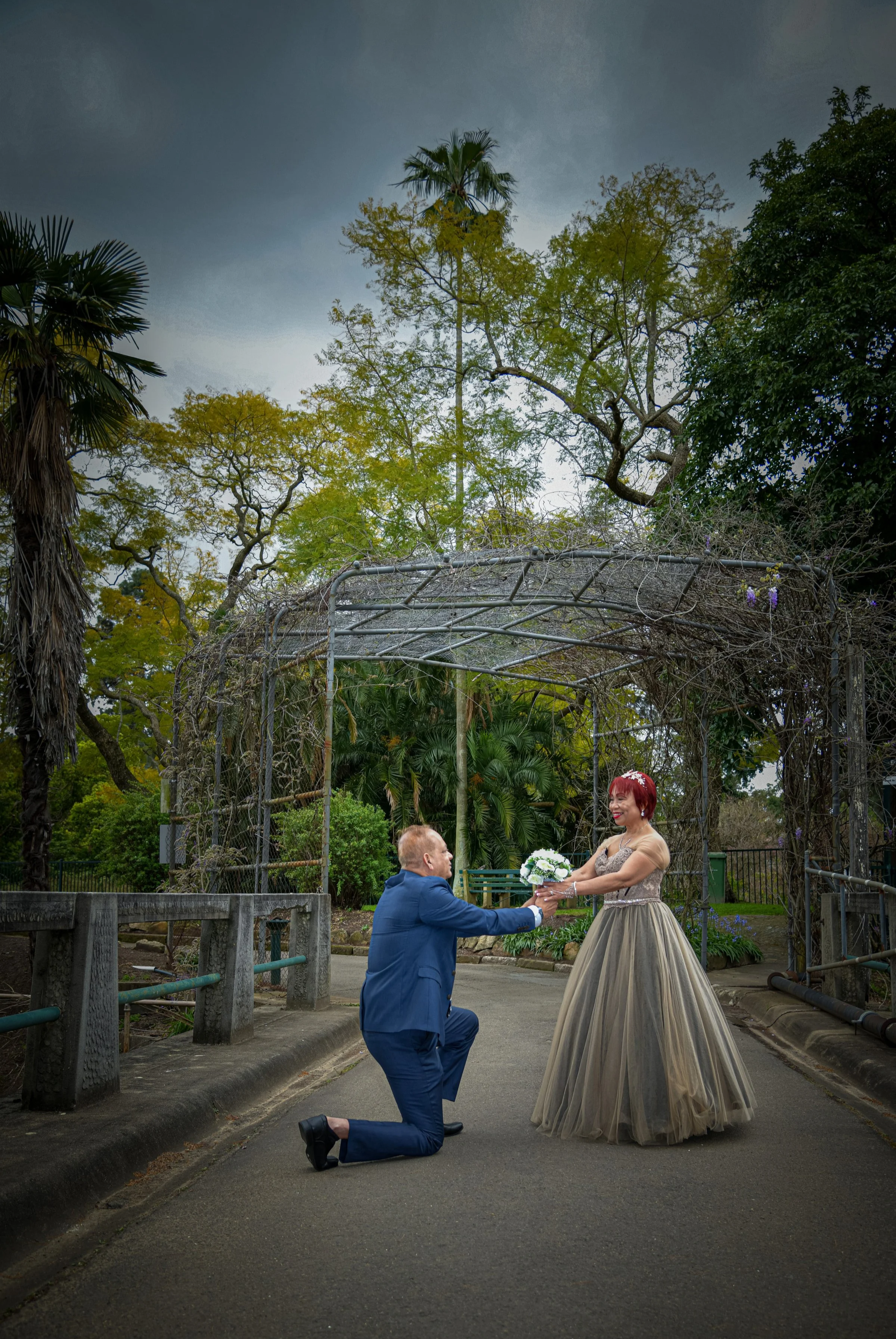 A man in a blue suit proposing marriage to a woman in a formal gown in a park.