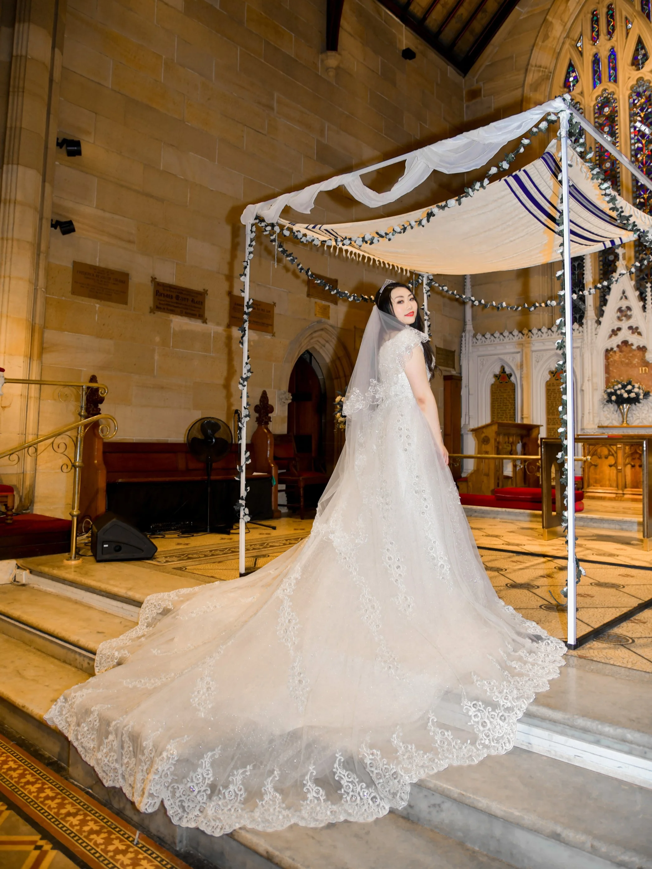 A bride in a white wedding gown with a long train stands inside a church under a canopy, with stained glass windows in the background.