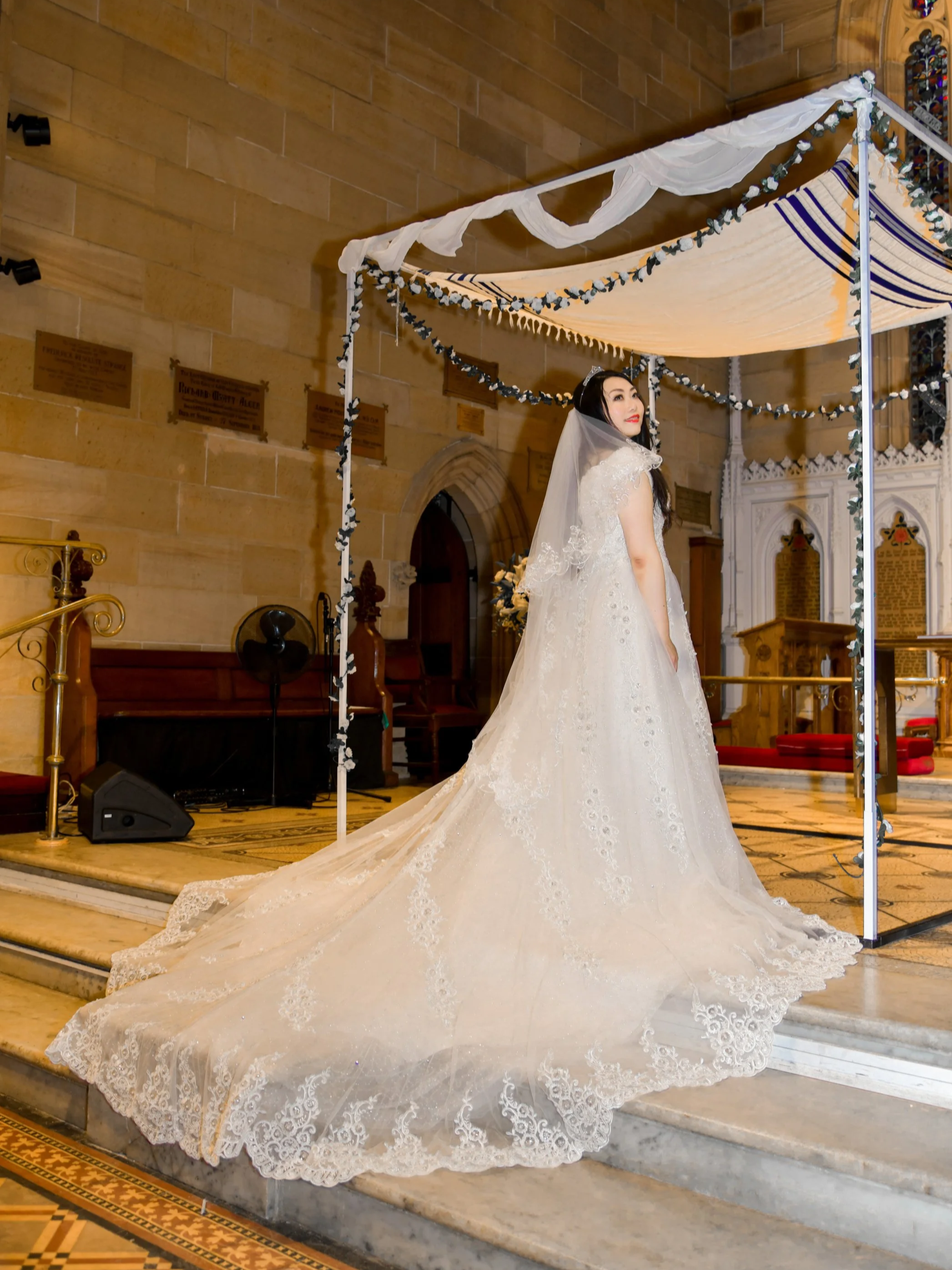 A bride in a white wedding gown with a long lace train standing inside a church, looking over her shoulder at the camera. The church features stone walls, wooden pews, and decorative plaques.