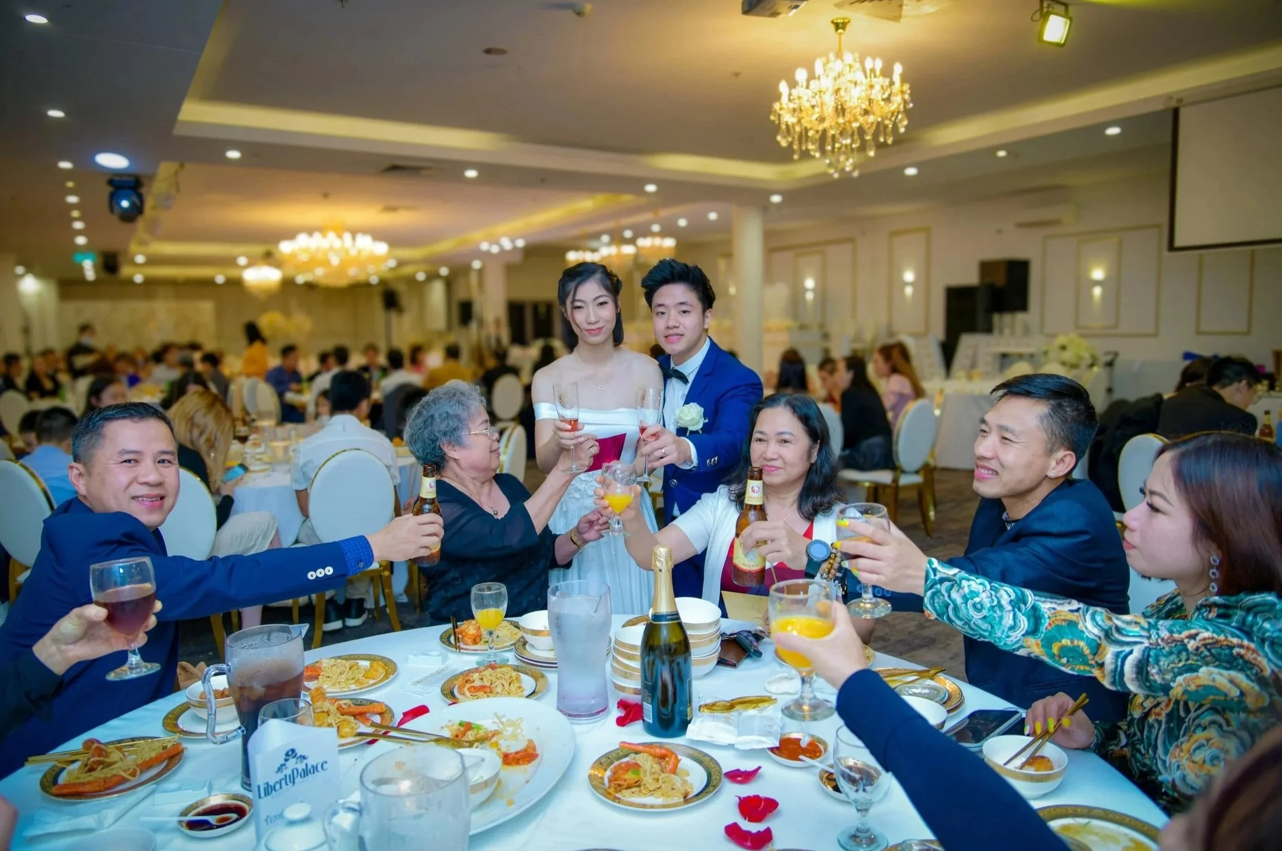 A group of people celebrating at a wedding reception, raising glasses in a toast around a table with food and drinks in a decorated banquet hall with chandeliers.