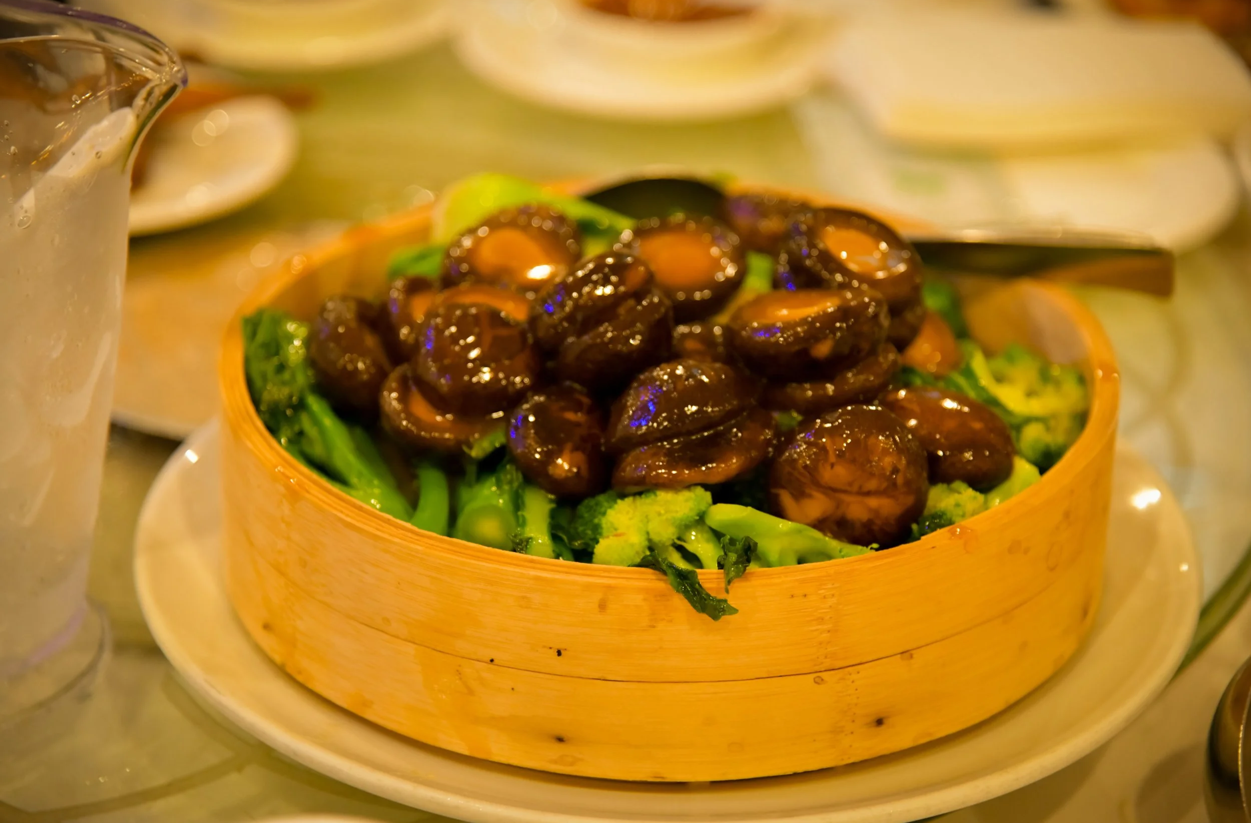 A bamboo steamer basket filled with cooked mushrooms on a bed of green vegetables, placed on a white plate with a spoon nearby, in a dining setting.