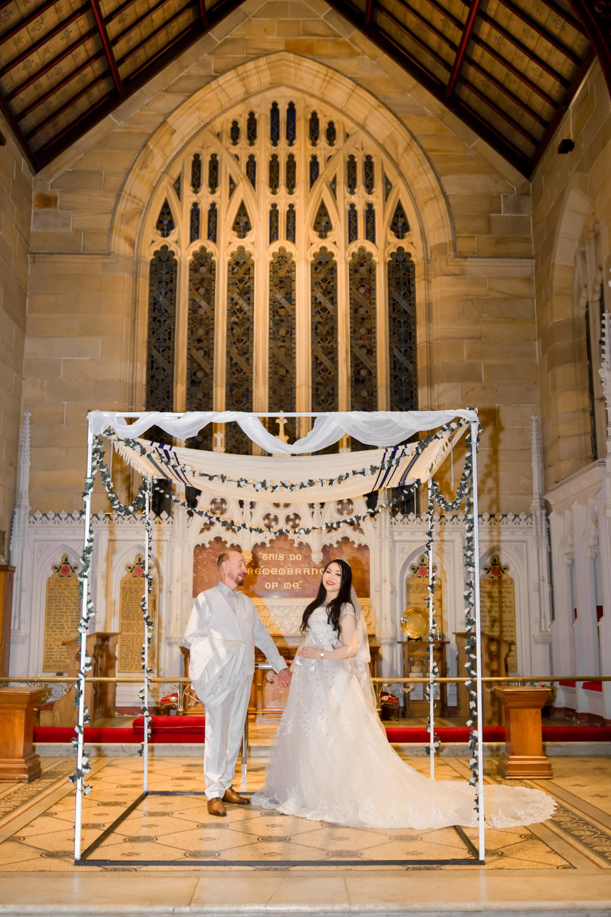 A bride and groom holding hands inside a church, standing under a decorated canopy, with a large stained glass window behind them and a sign that says 'This do remember of me' in the background.